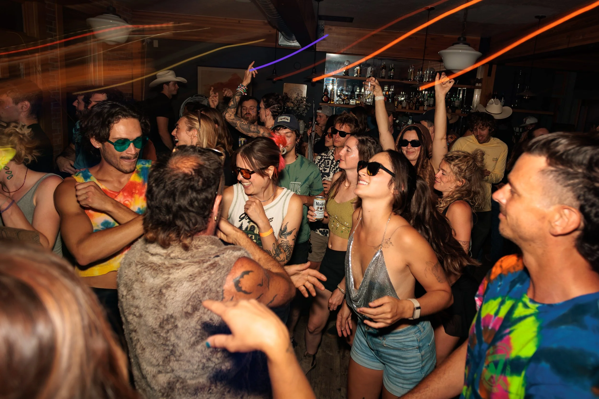 People dancing and enjoying a lively party in a crowded indoor venue with warm lighting, some wearing sunglasses, themed clothing, and holding drinks, with light streaks indicating dancing and movement.