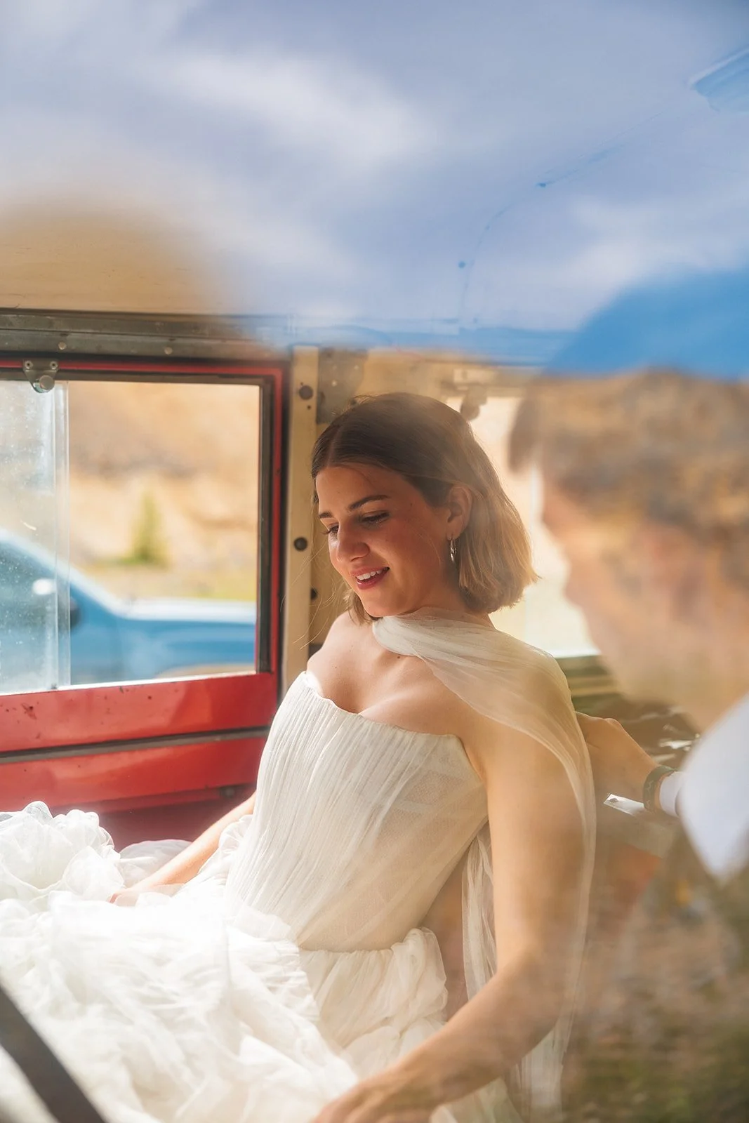 A woman in a white dress sitting inside a colorful vintage vehicle, smiling and looking down with a background of blue sky and rocky landscape.