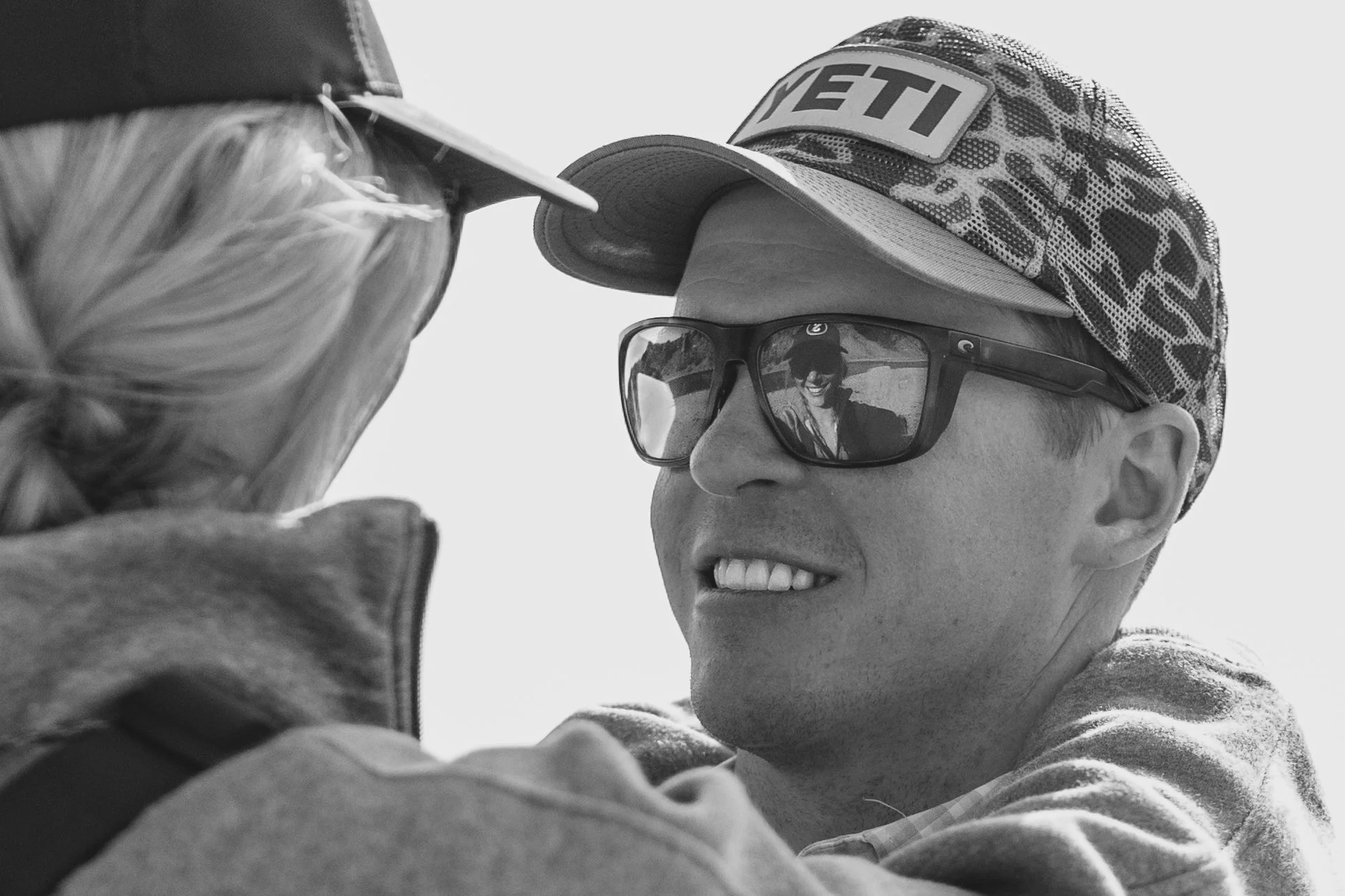 A person wearing a camo-patterned baseball cap and black sunglasses is smiling and taking a selfie with an older woman with blonde hair. Both are outdoors in bright light.