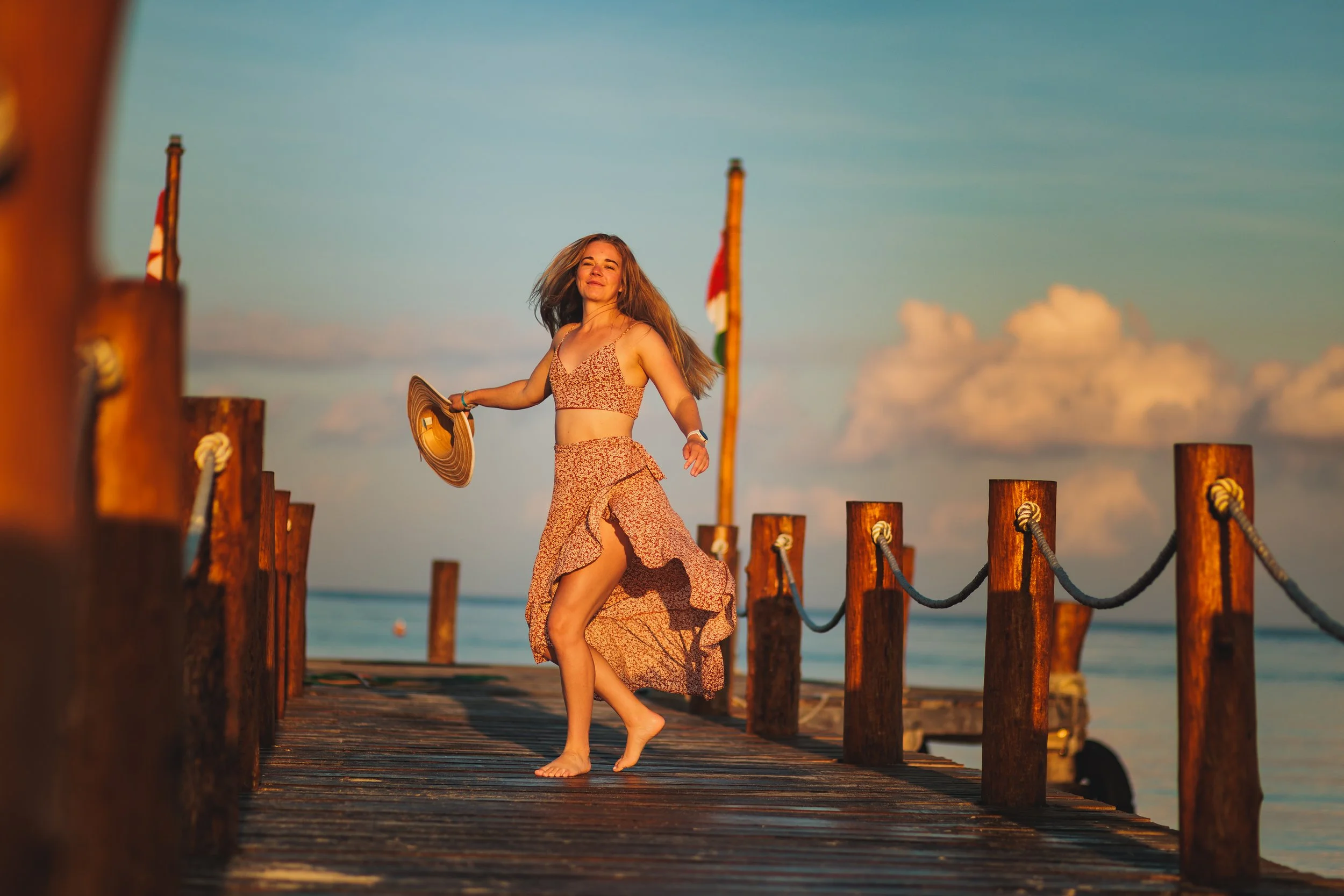 A woman in a floral summer dress with a slit, holding a straw hat, dancing on a wooden pier at sunset with the ocean in the background.