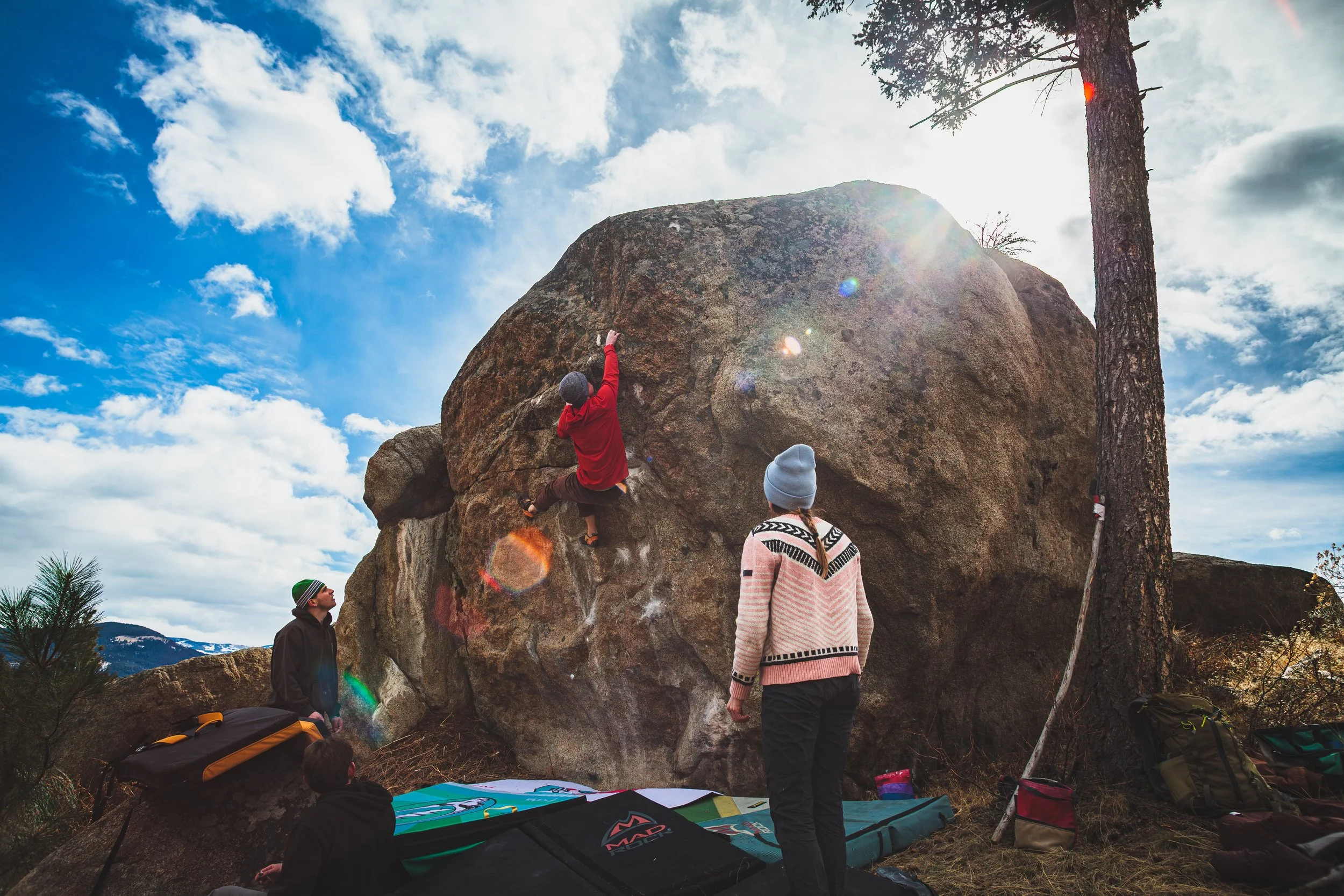 A person is bouldering on a large rock outdoors beneath a partly cloudy sky, with others watching nearby.