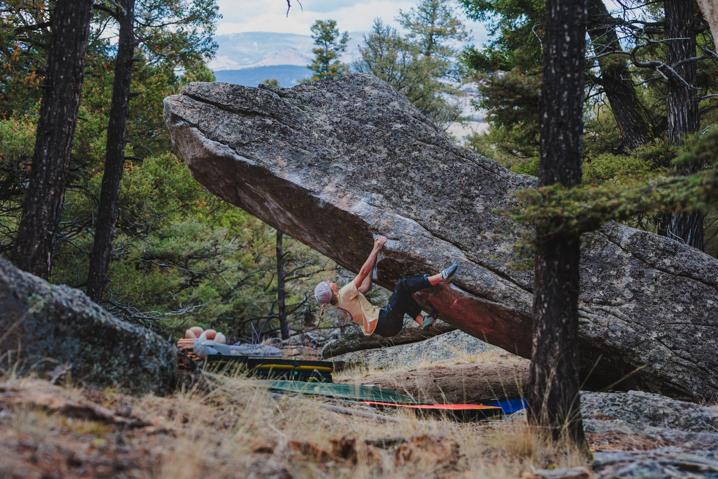 A woman rock climbing on a large overhanging boulder in a forested area with trees and mountains in the background.