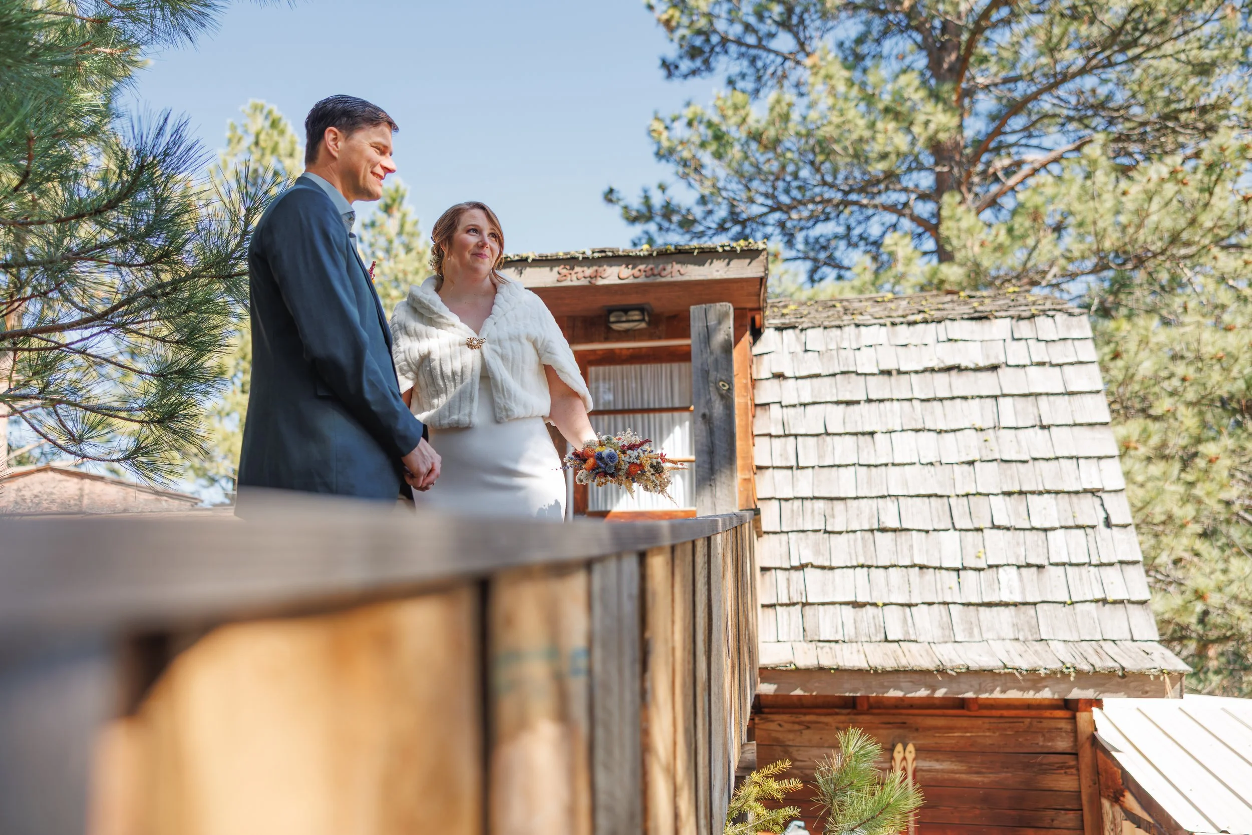 A man and woman dressed in formal attire holding hands and smiling outdoors during a wedding ceremony with a wooden backdrop and surrounded by trees and blue sky.
