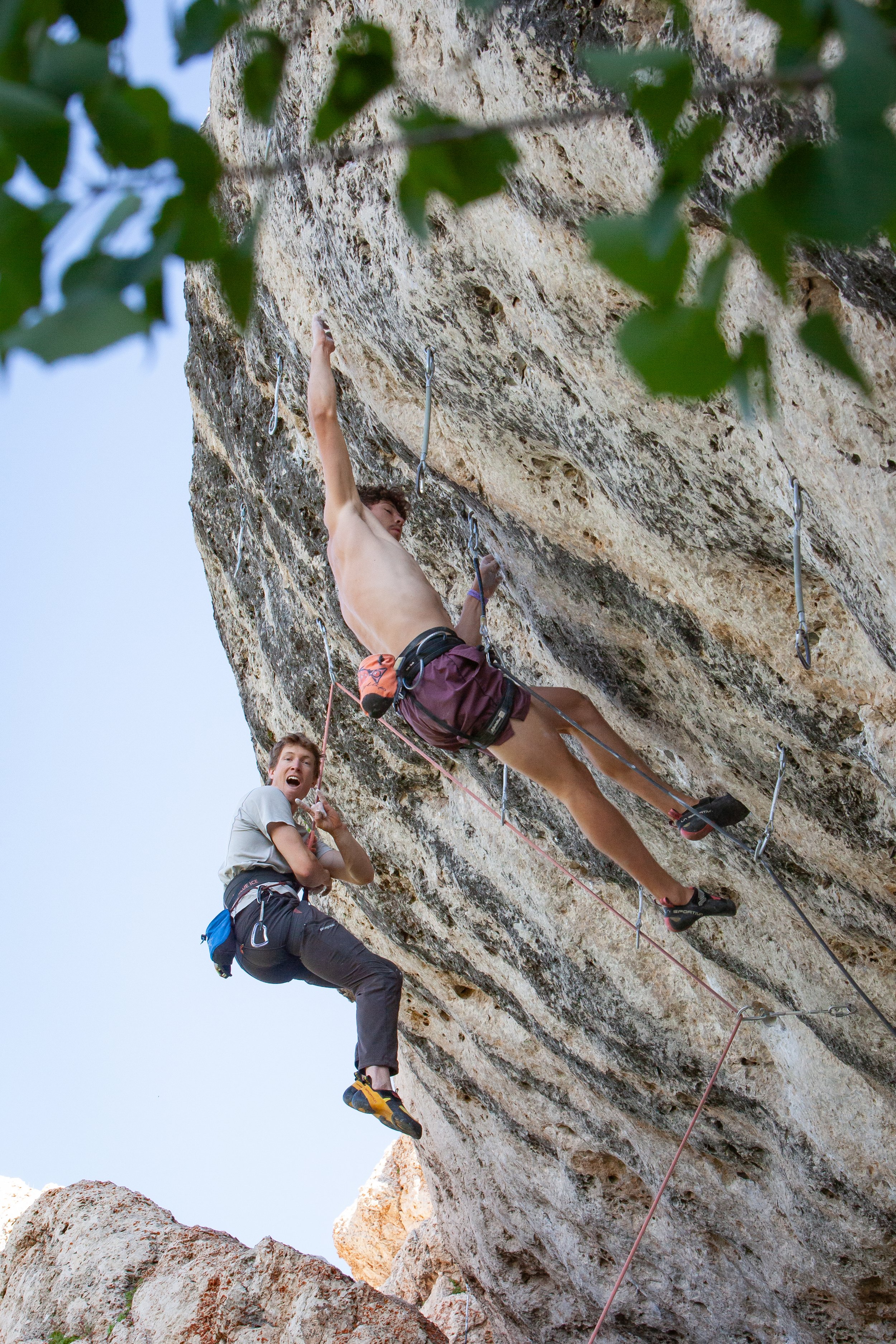 A man and woman rock climbing on a steep outdoor cliff, with the man reaching for a handhold and the woman looking surprised or excited beneath him, surrounded by greenery.