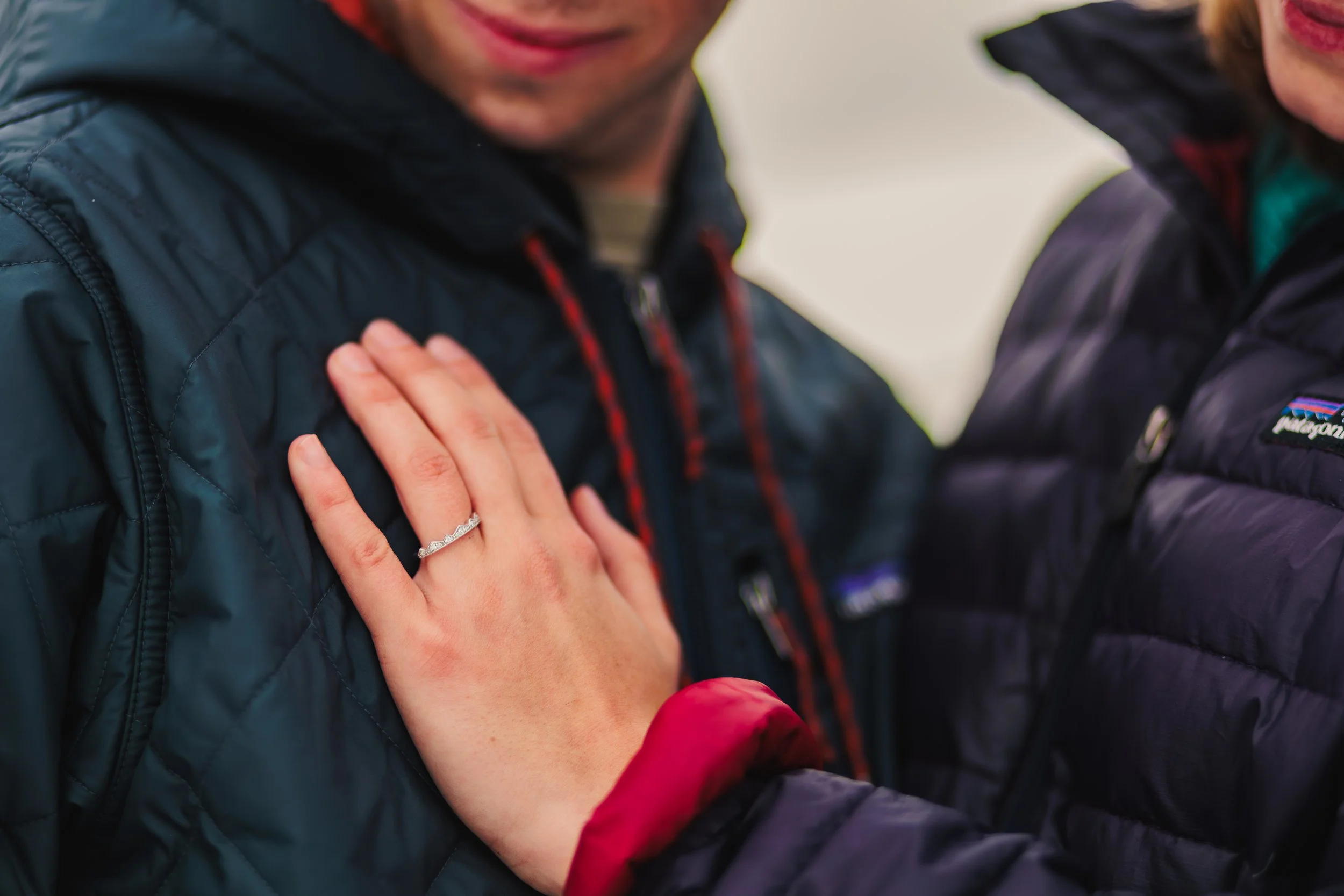 A woman wearing a dark jacket with a diamond ring on her finger is placing her hand on a man's chest. The man's face is partially visible, and he is wearing a navy blue jacket with a red zipper.