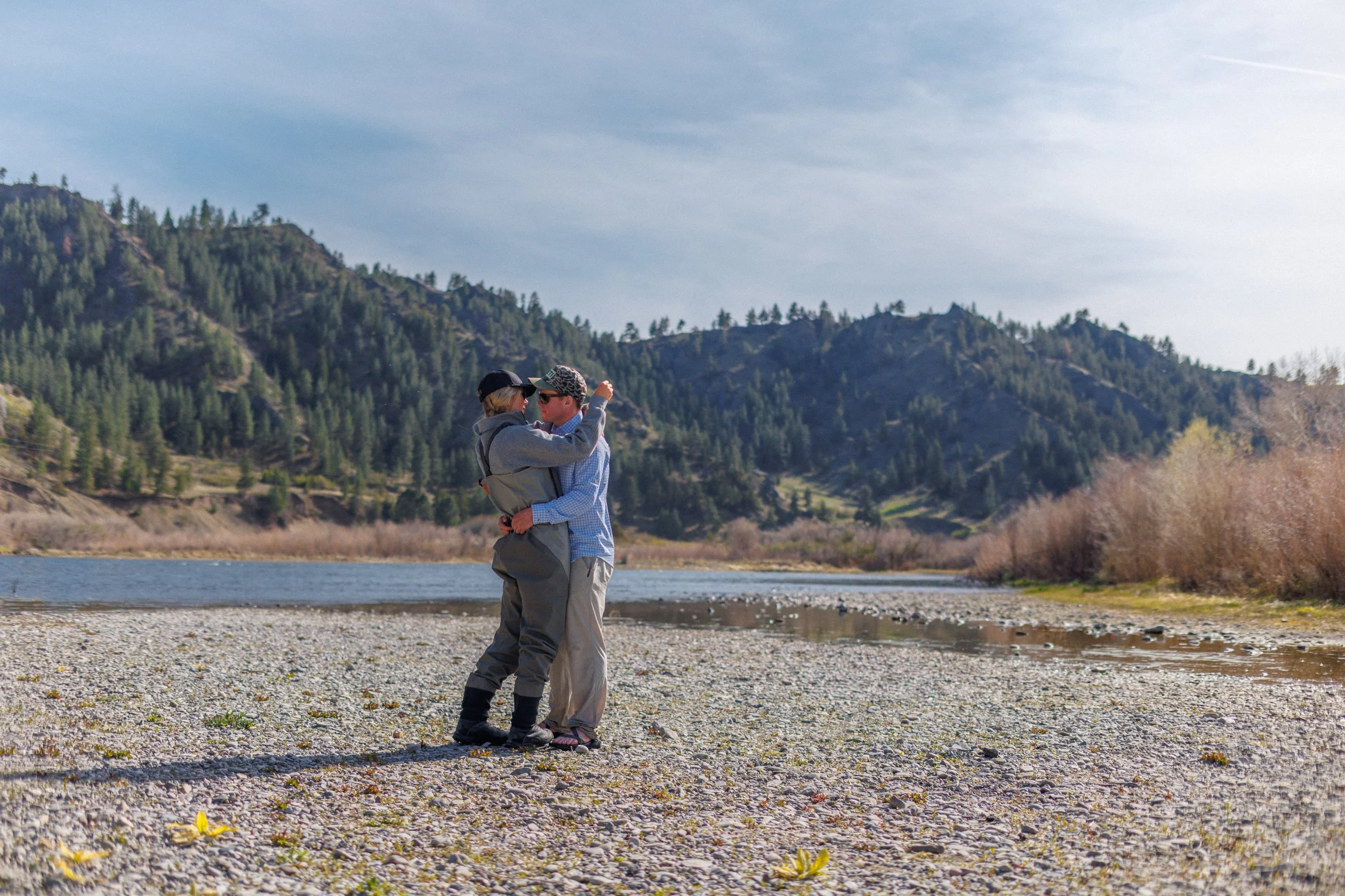 A couple standing on a rocky shoreline by a body of water, embracing each other, with mountains covered in trees in the background and a partly cloudy sky above.