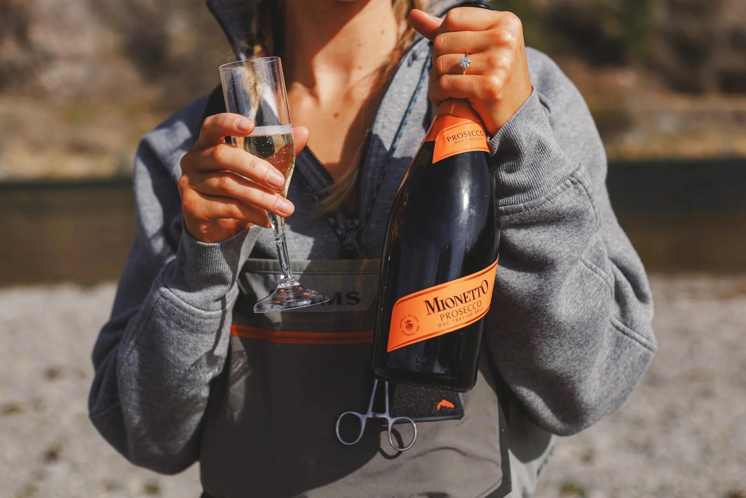 Person holding a glass of sparkling wine or champagne and a bottle of Mionetto Prosecco outdoors during daytime.