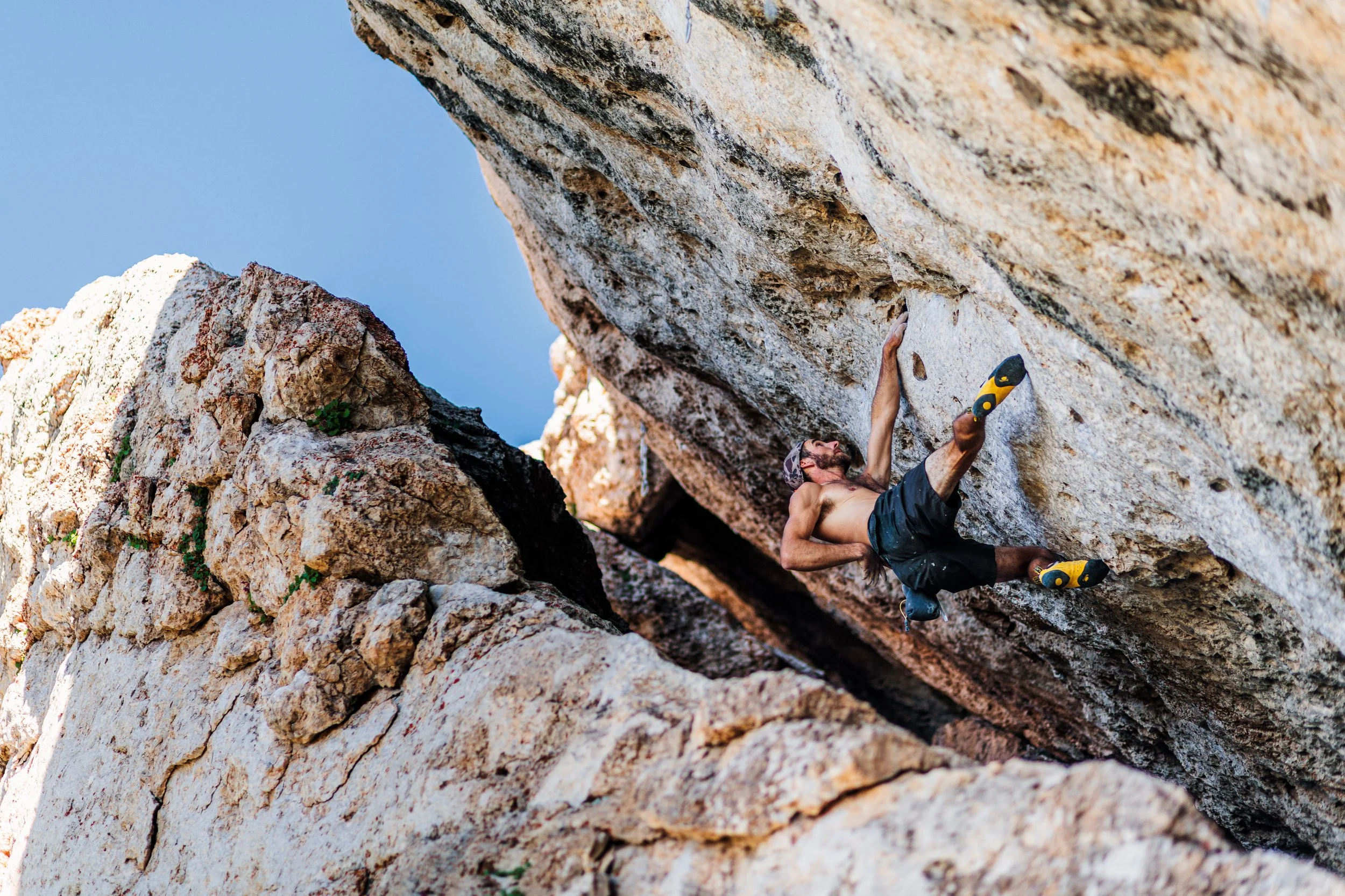 A shirtless man with a beard and a backwards cap rock climbing on a steep granite rock face in an outdoor setting with blue sky.