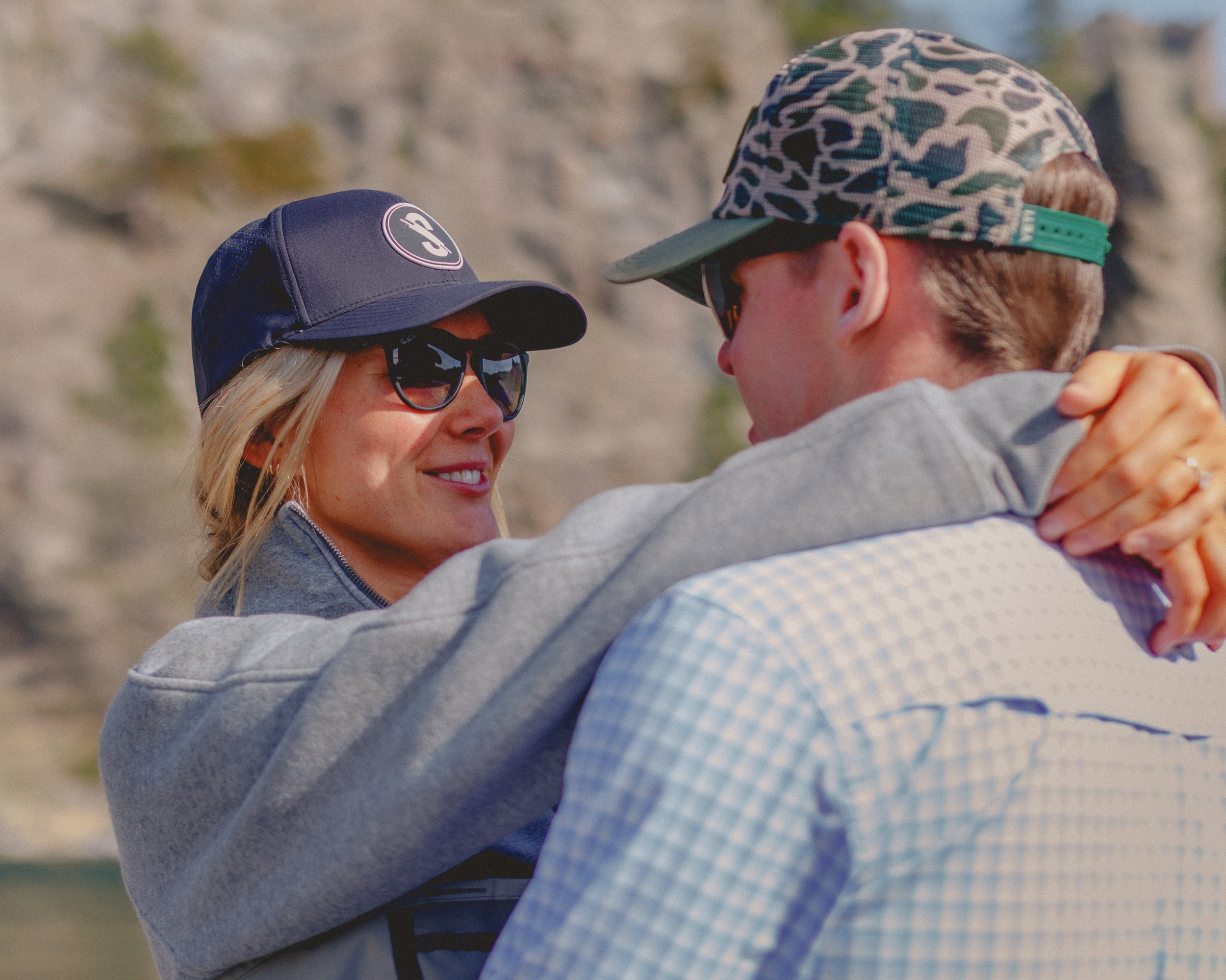 A couple, wearing hats and sunglasses, dancing outdoors with a rocky landscape in the background.