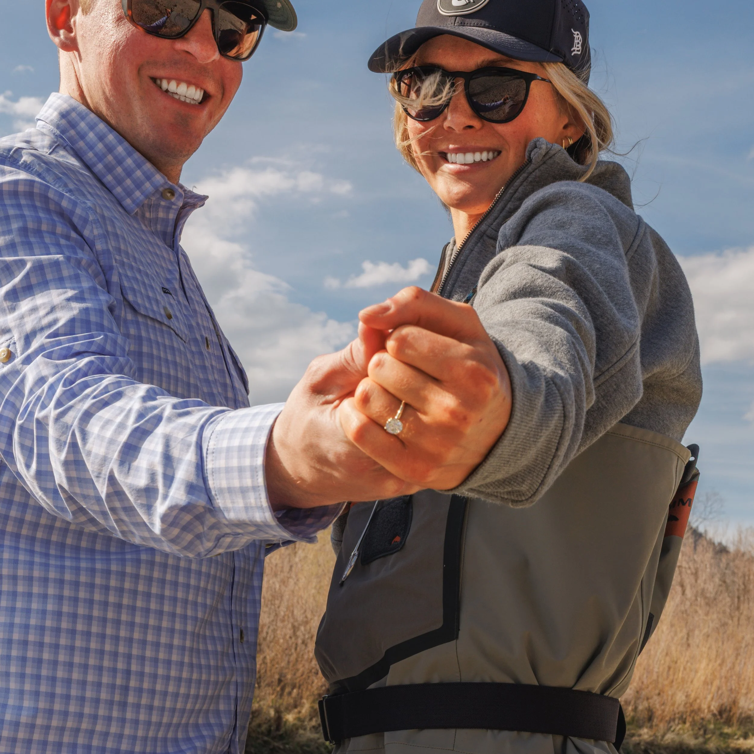 Close-up of a smiling man and woman standing outdoors on a sunny day, wearing sunglasses and hats, holding hands, with a blue sky and sparse clouds in the background.