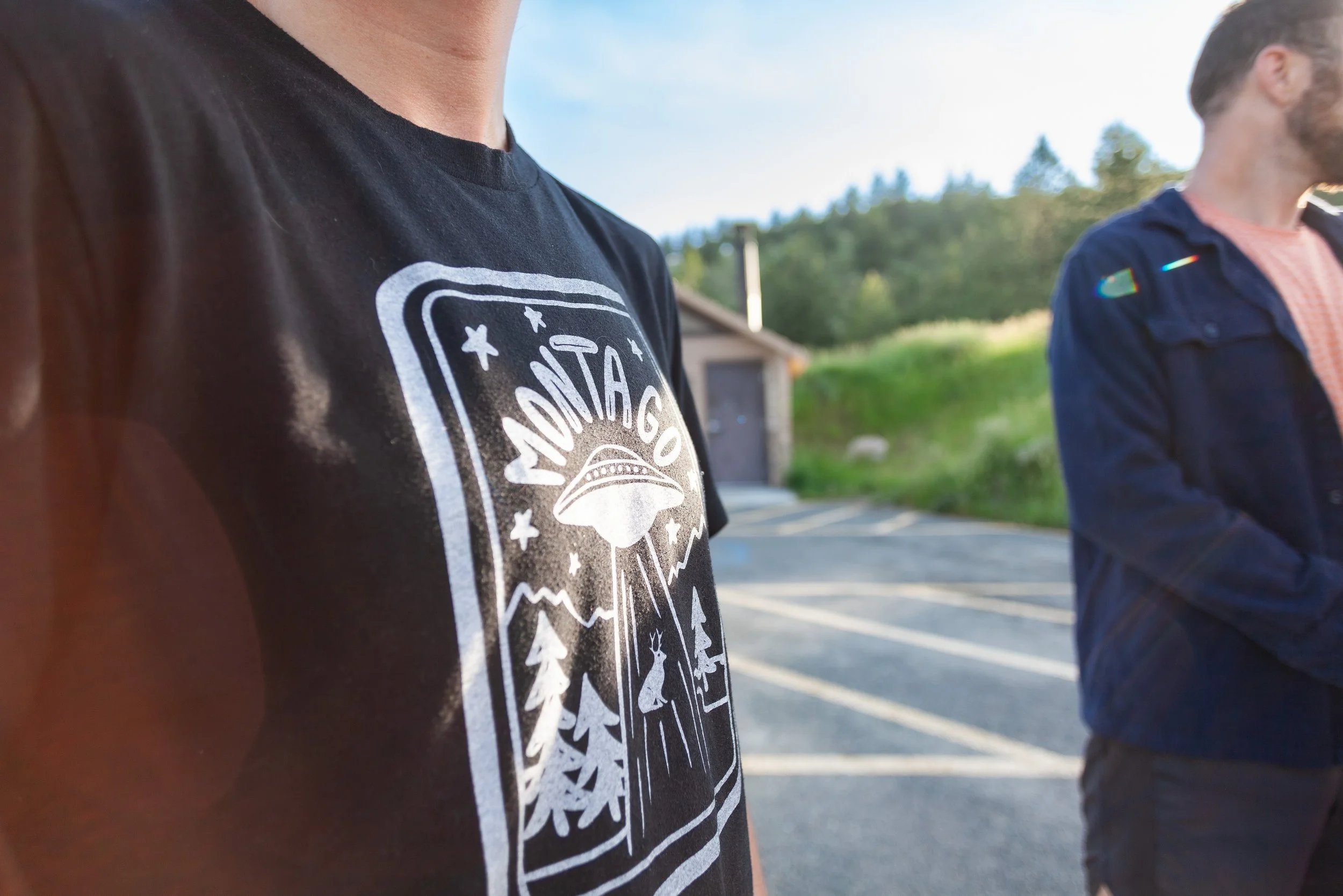 Close-up of a person's black t-shirt featuring a graphic with a UFO, the word 'Montago', mountains, trees, a deer, and stars. Another person is standing nearby in an outdoor parking lot with a building and greenery in the background.