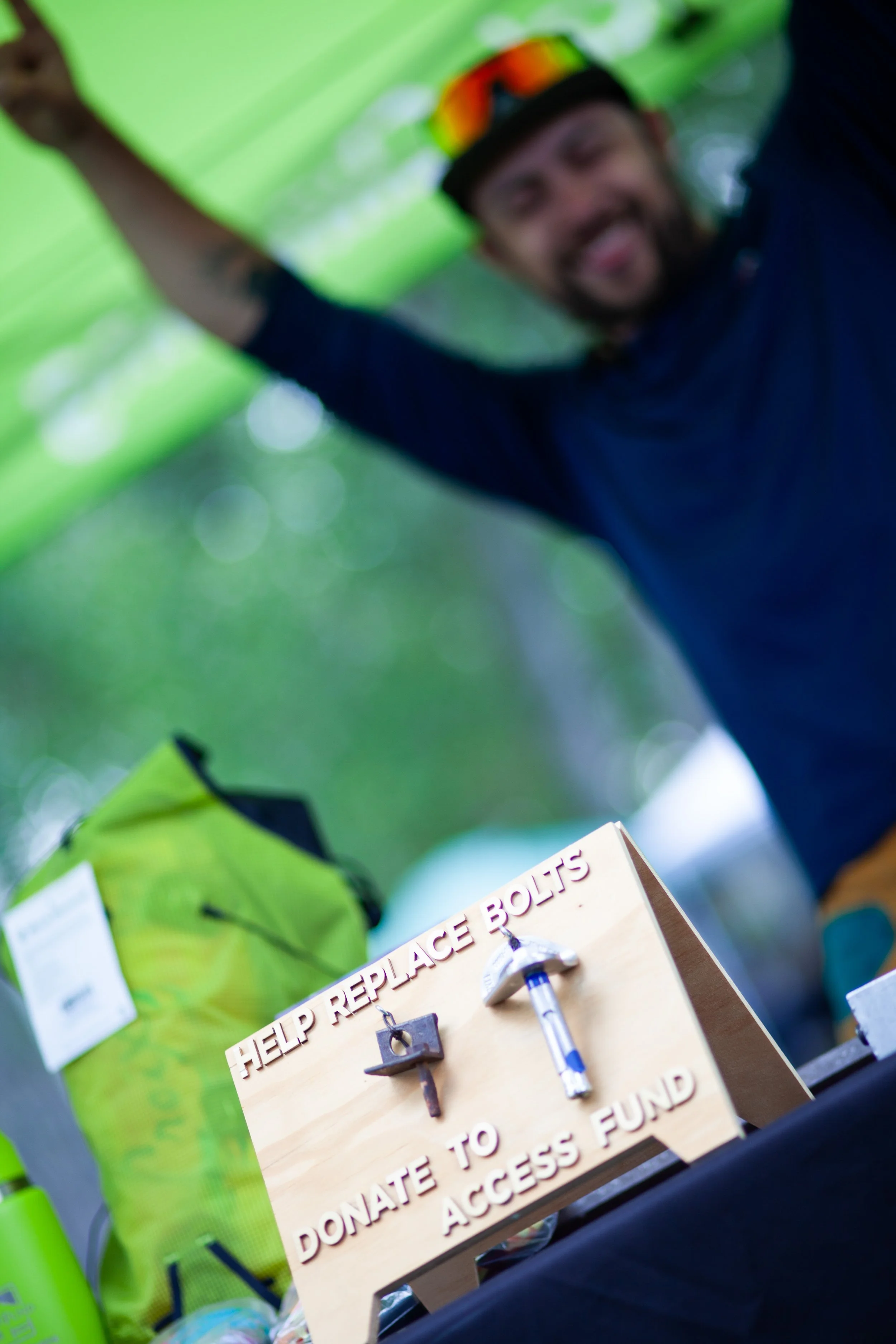 A man in a black shirt and a helmet smiling, standing outdoors under trees. In the foreground, there is a wooden donation box with a sign reading 'Help Replace Bolts' and 'Donate to Access Fund', along with a key and a screwdriver.
