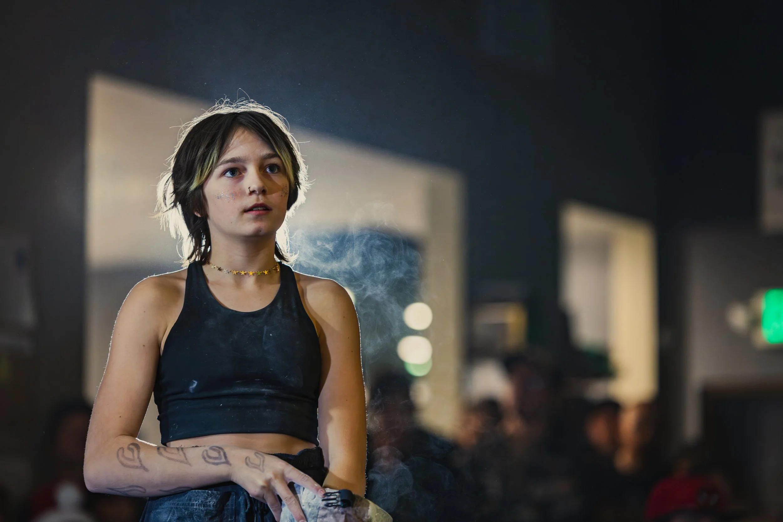 A young woman with short dark hair, tattoos on her arm, wearing a black tank top and a star necklace, stands indoors with a slightly surprised or curious expression, with smoke around her and blurred people in the background.