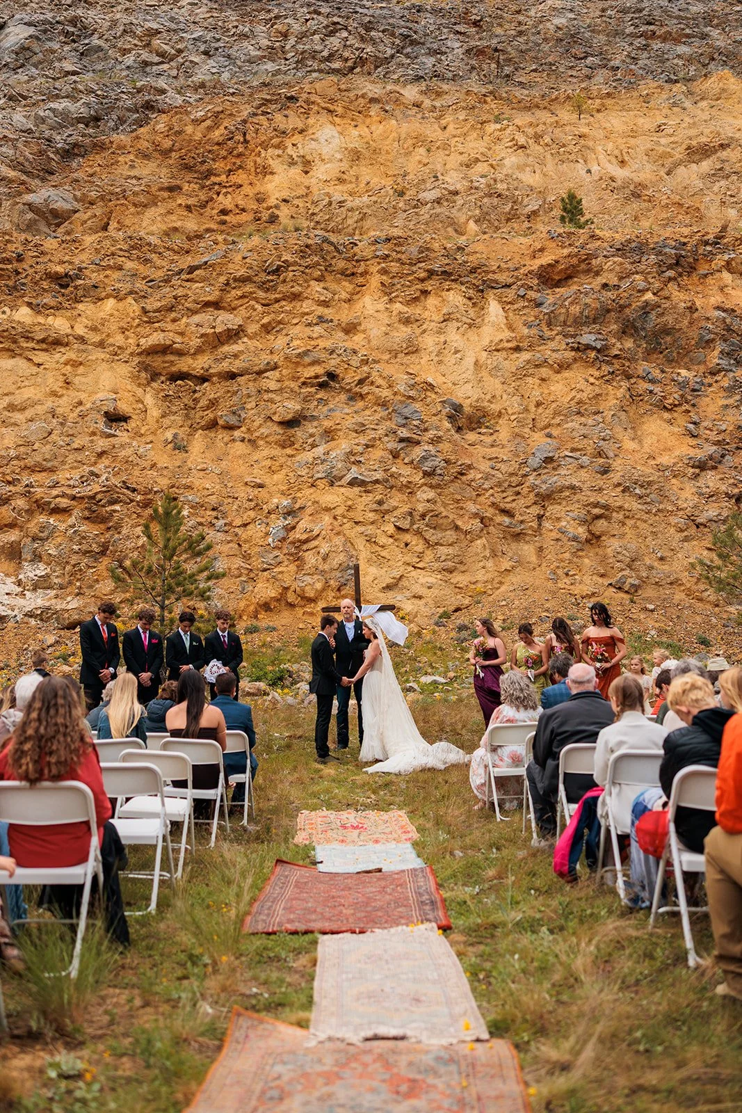 A wedding ceremony taking place outdoors at the base of a rocky cliff. The bride and groom are exchanging vows, surrounded by bridesmaids and groomsmen, with guests seated in white chairs.