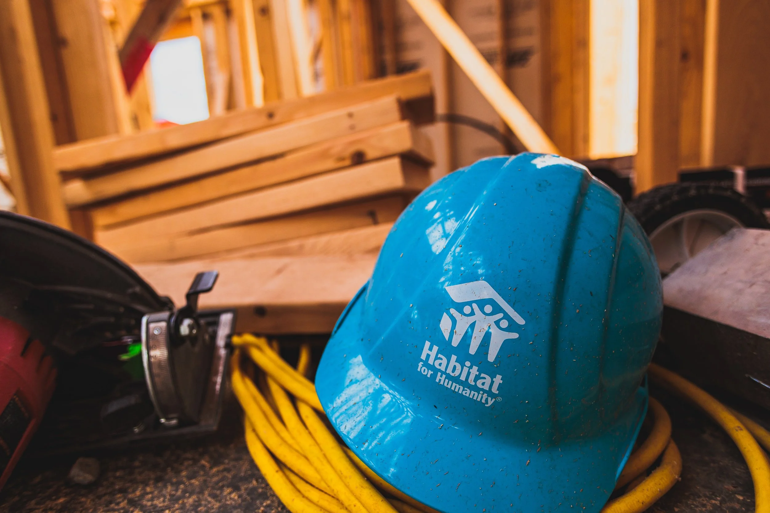 A blue hard hat with the Habitat for Humanity logo resting among construction tools and yellow extension cords inside a building under construction.