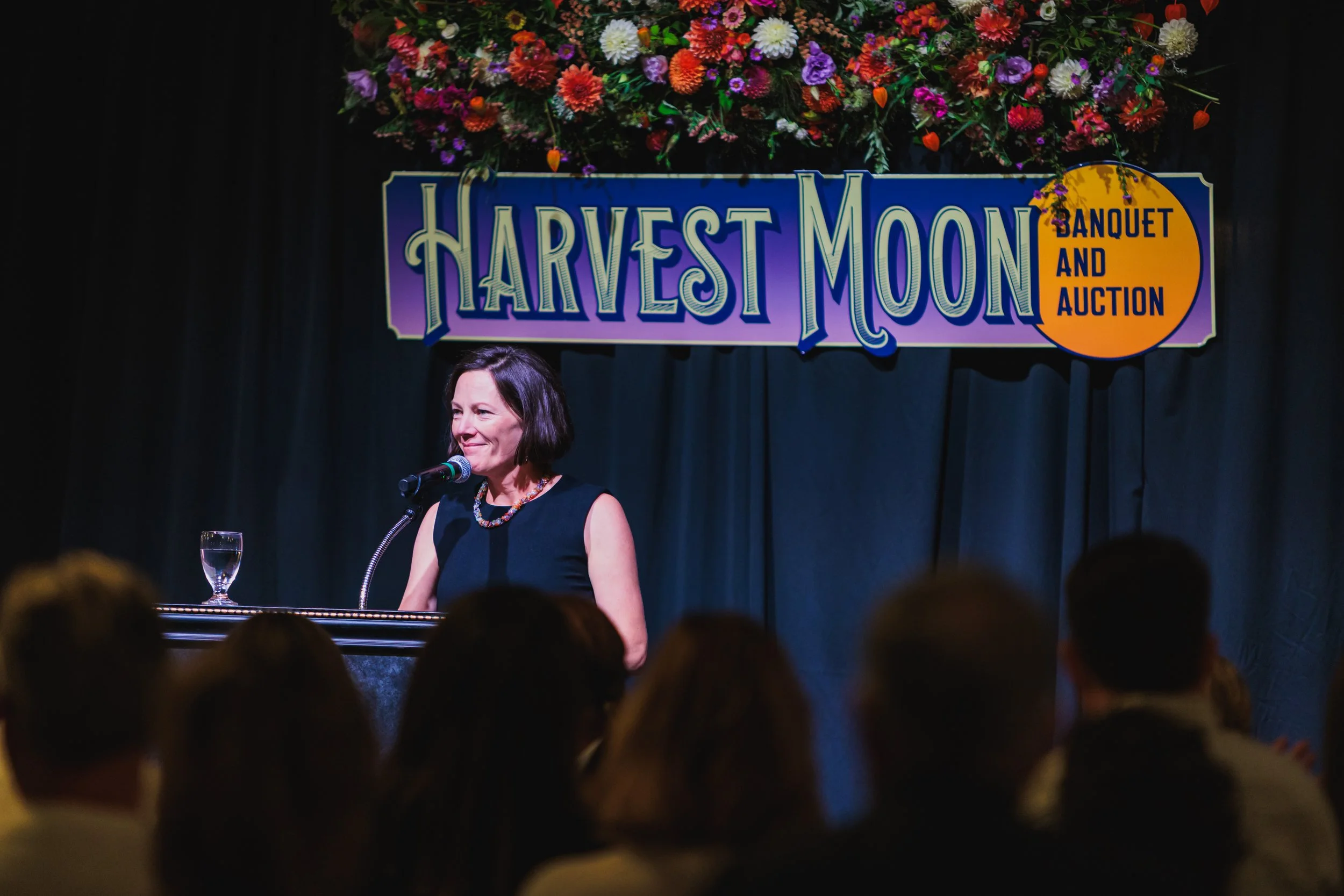 A woman with dark, shoulder-length hair speaking at a podium during an event called 'Harvest Moon Banquet and Auction,' with a glass of water on the podium and an audience in front. Behind her is a floral arrangement and a sign with colorful letterin