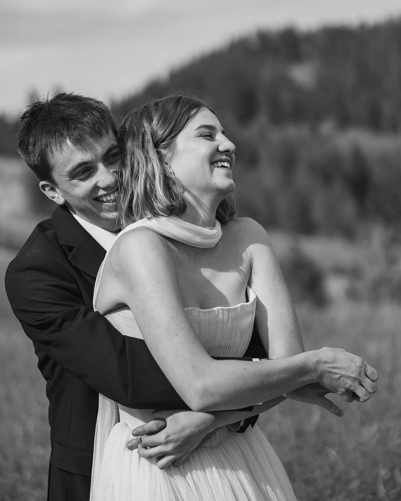 Black and white photo of a smiling couple outdoors, with the man embracing the woman from behind. The woman is looking to her right, and they both appear happy, laughing. The background shows trees and a body of water.