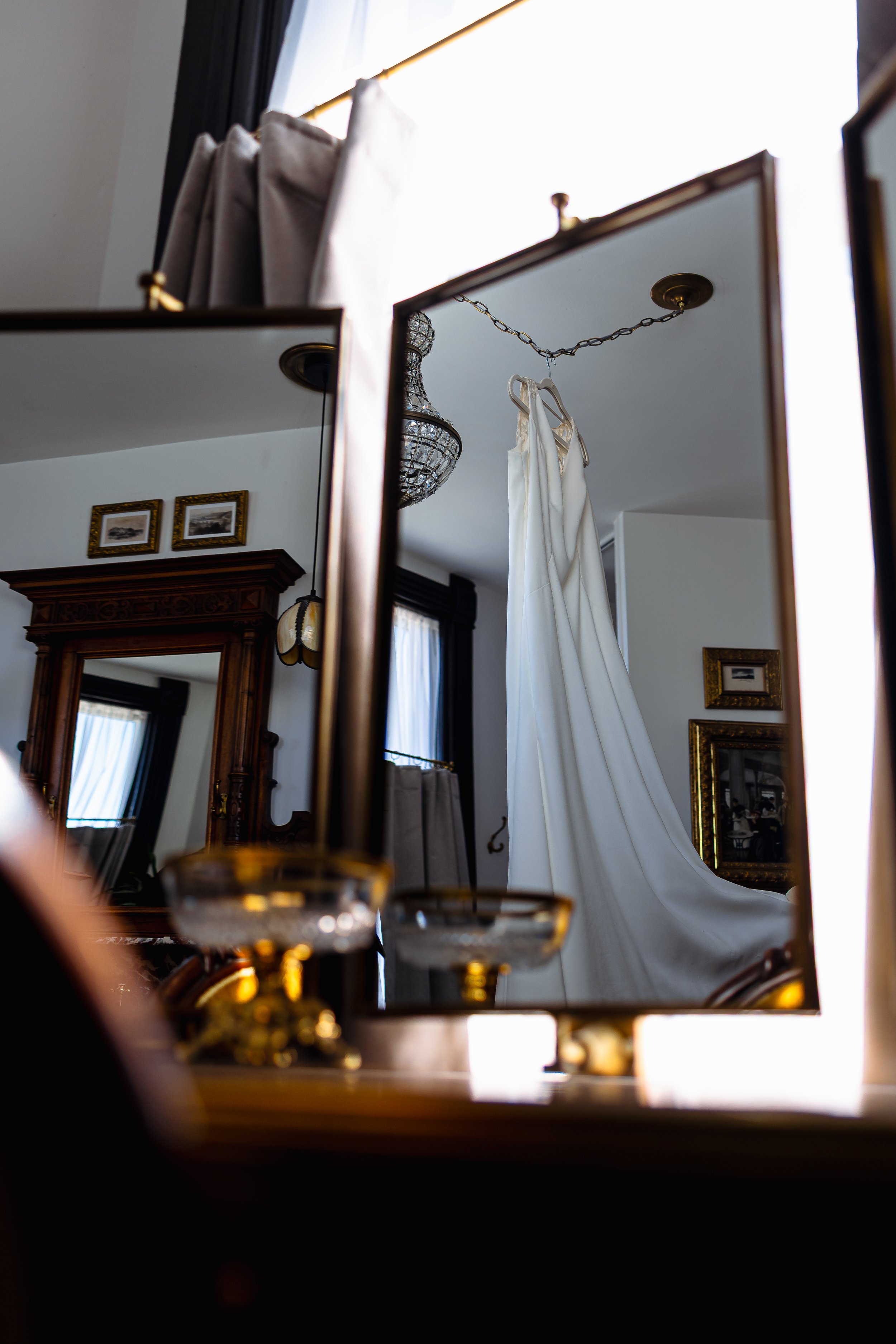 A reflection mirror shows a room with a white dress hanging from a chain hook, an ornate chandelier, framed pictures on the wall, and a wooden cabinet. The room has natural light coming from a window.