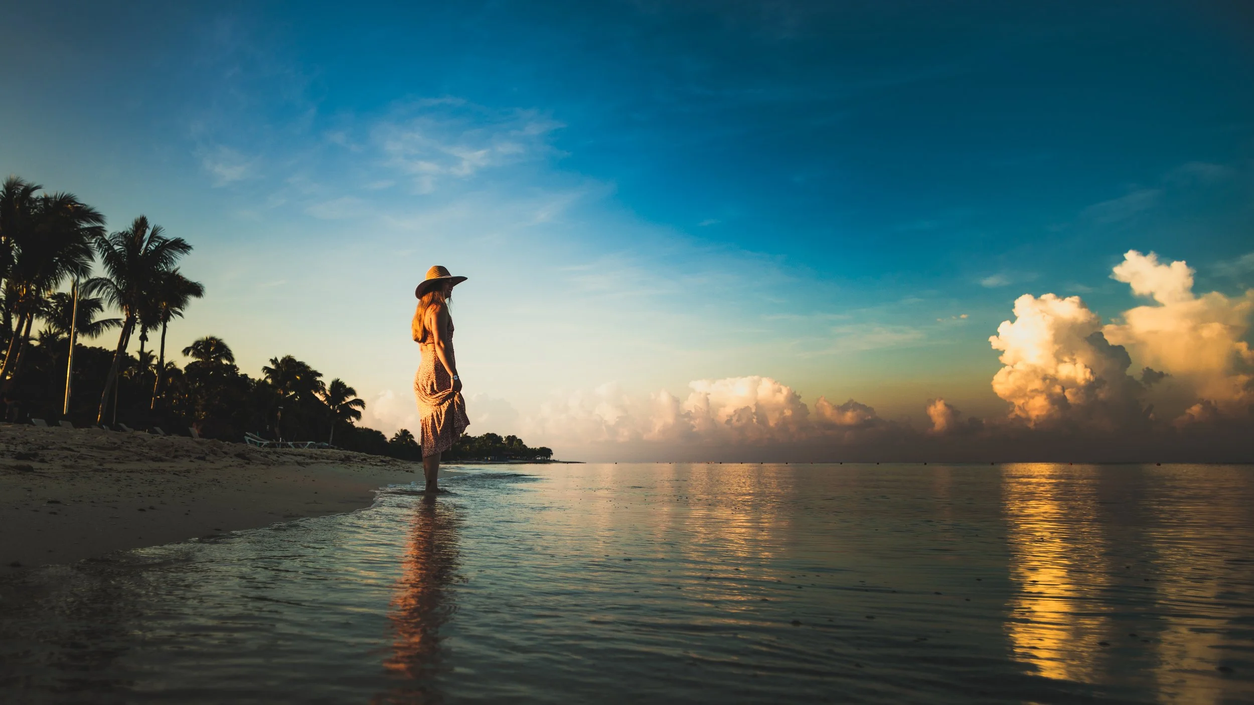 A woman standing in shallow water along a beach at sunset, with palm trees on the left and clouds on the horizon.