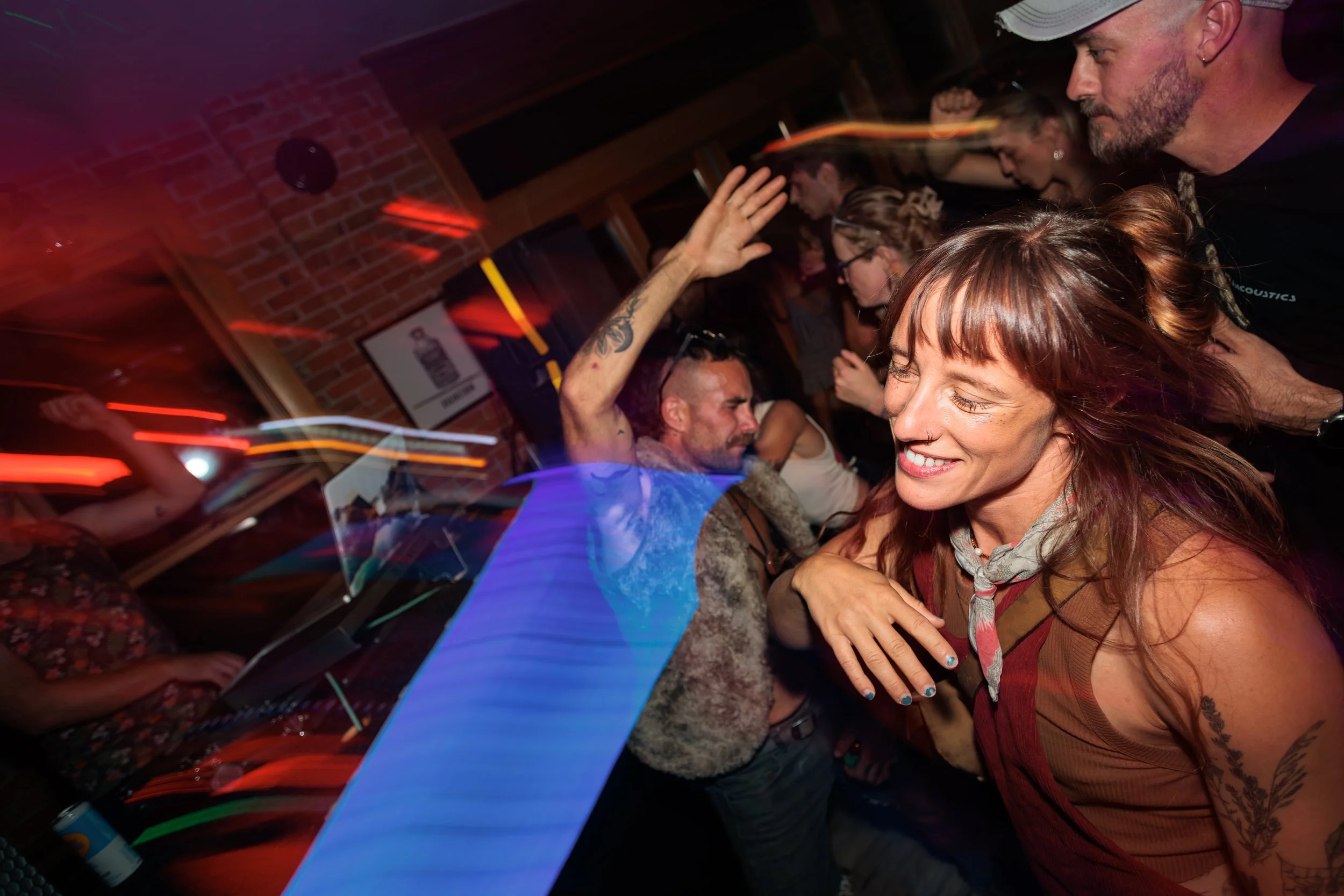 People dancing and socializing in a dimly lit indoor venue with colorful and blurry light streaks, exposed brick wall, and drinks on the table.