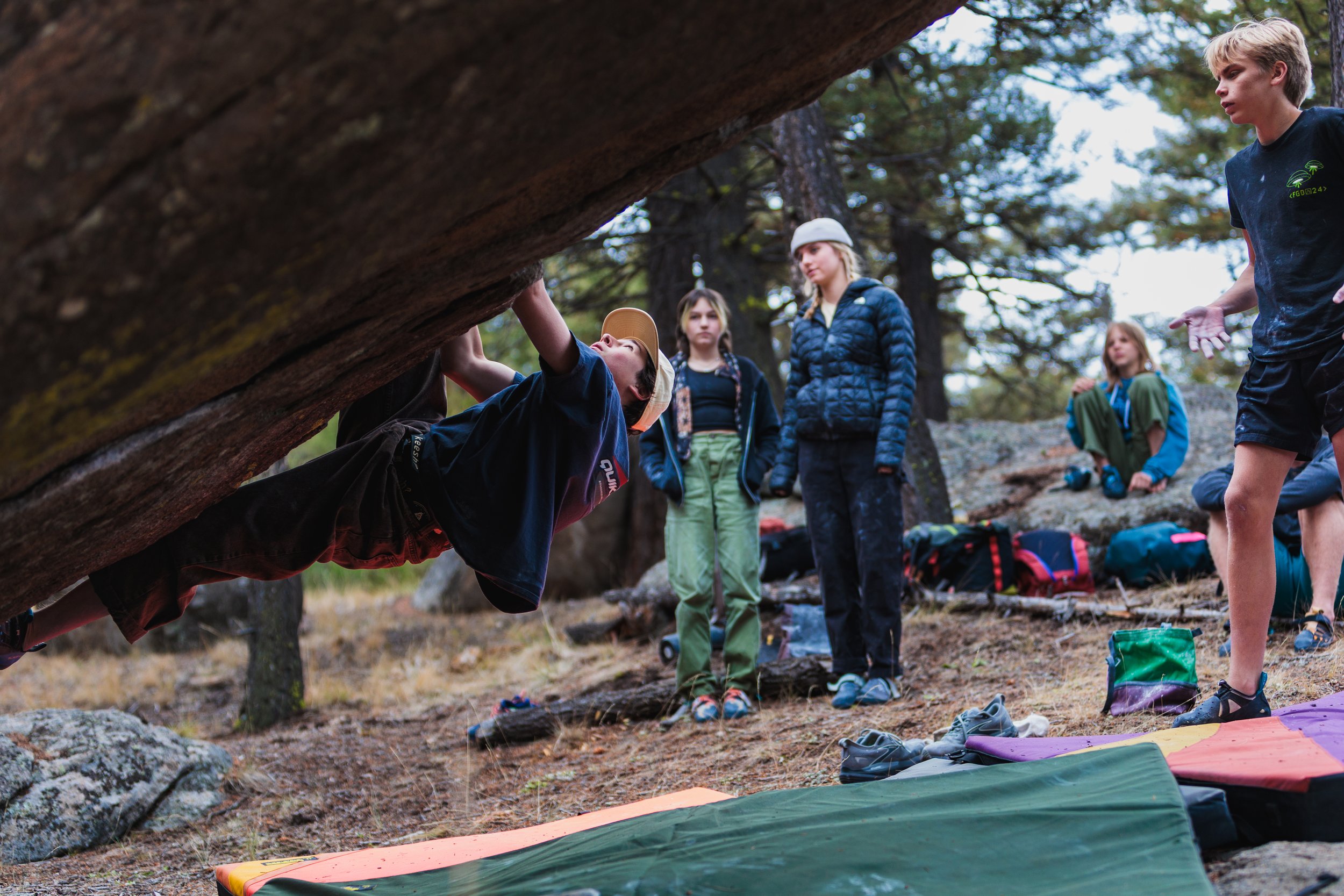 A group of young people outdoors in a forest, with one boy bouldering under a large rock while others watch.