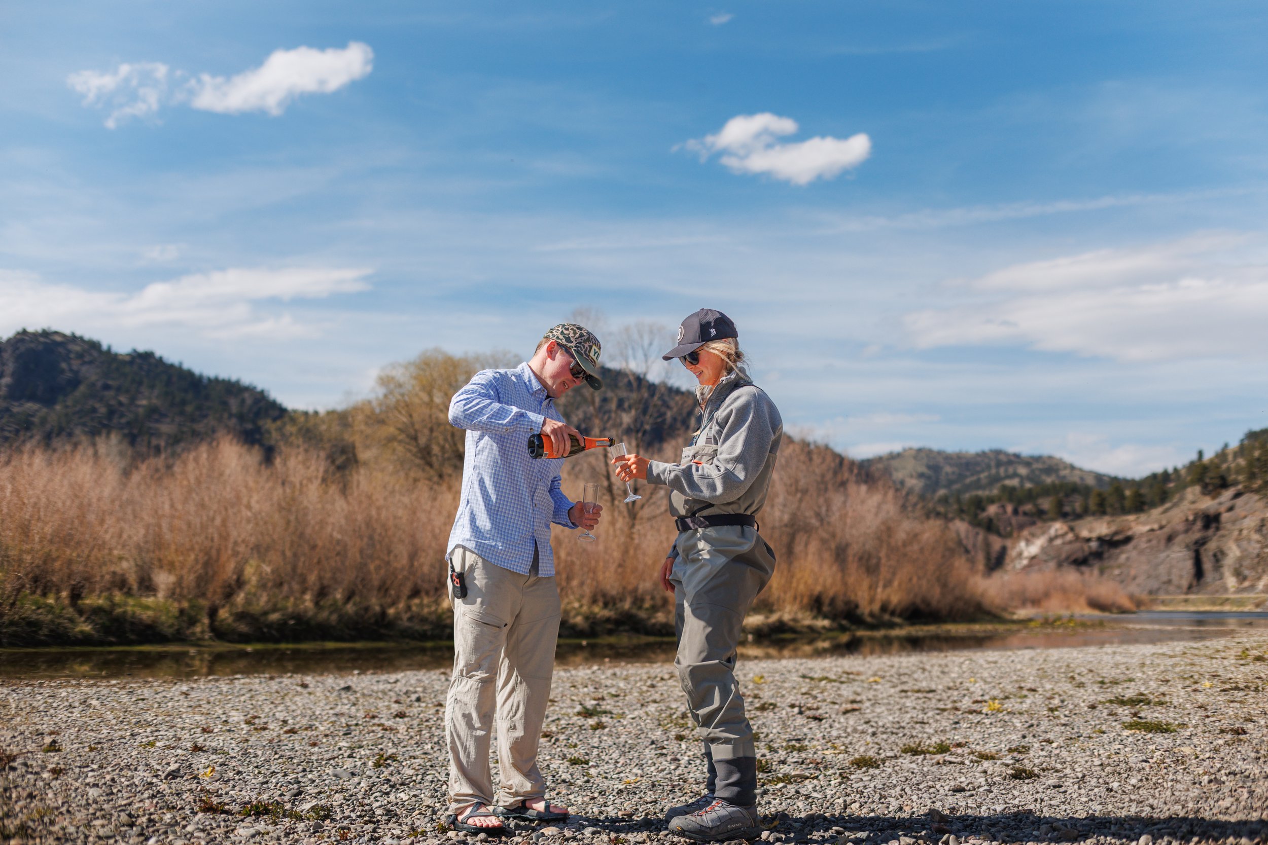 Two people standing outdoors on a rocky riverbank, one pouring a drink into a glass held by the other, with hills and trees in the background under a partly cloudy sky.