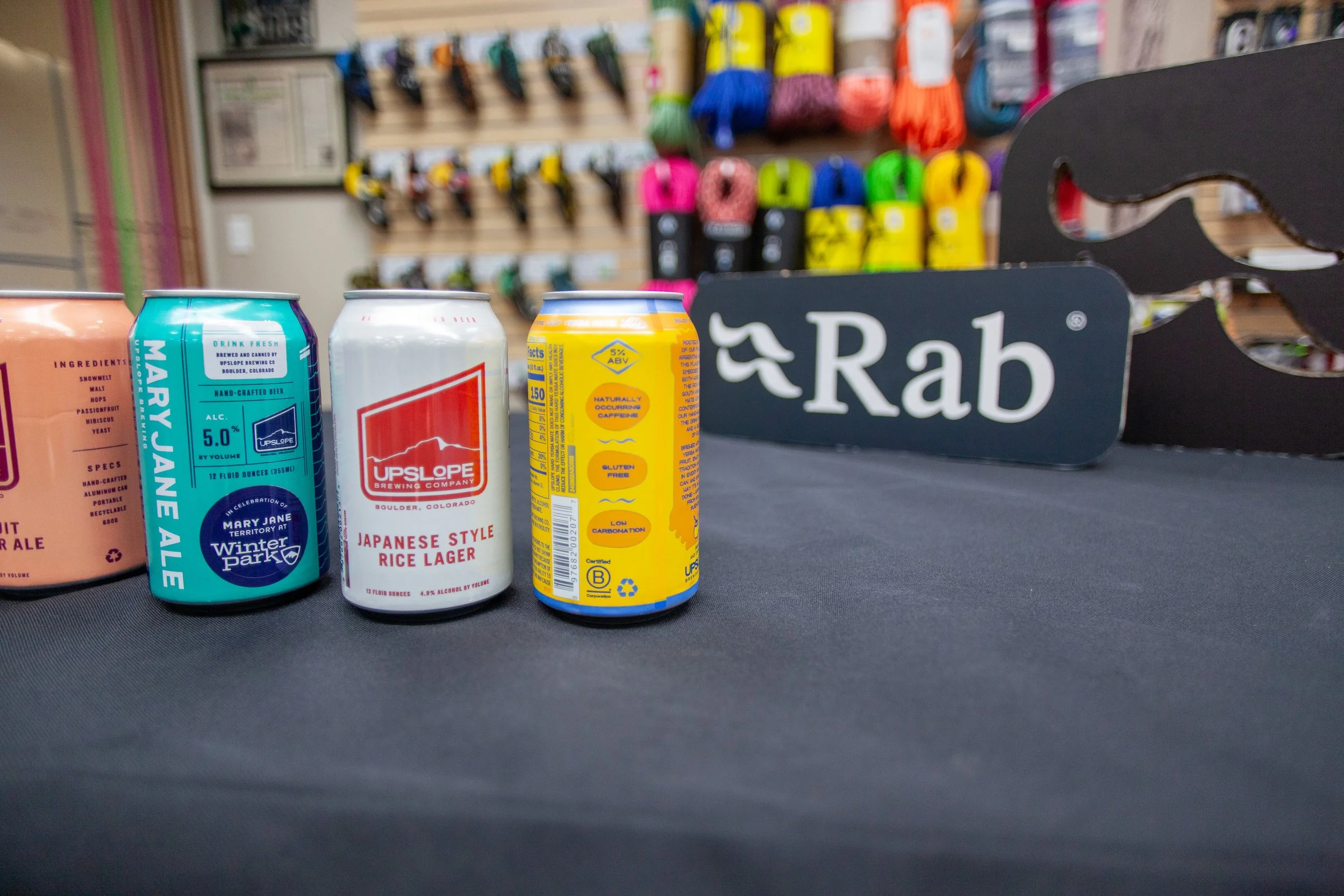Three cans of beverages on a black table, with a background of colorful rope spools and a Rab sign.