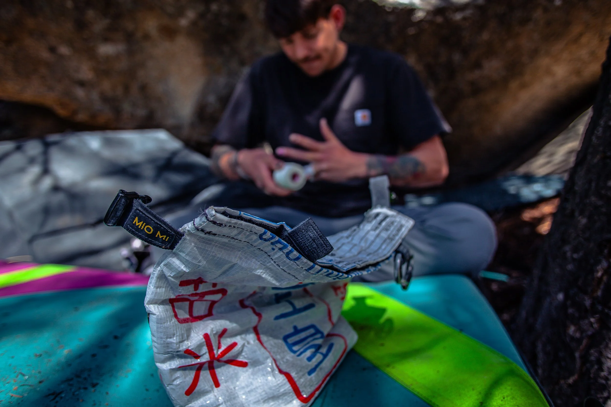A man sitting outdoors on a rock surface, preparing for an outdoor activity, with gear including a brightly colored surfboard and a dry bag with red and blue writing.