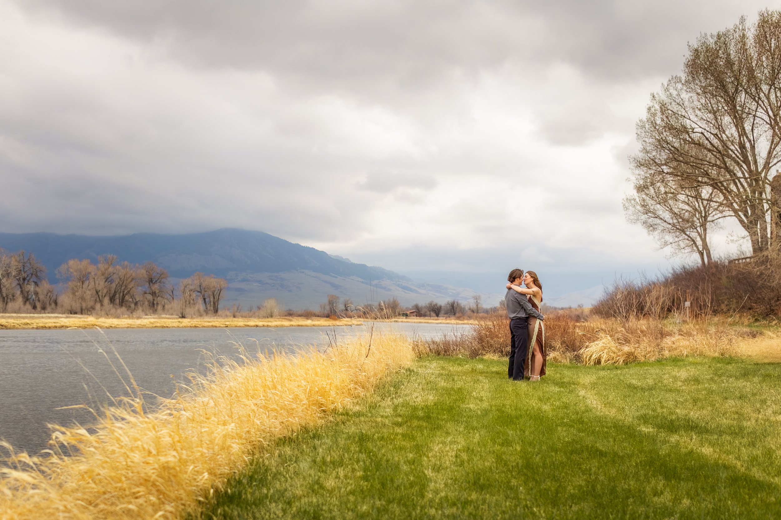 A couple embracing near a body of water in a scenic landscape with mountains, leafless trees, and cloudy sky.