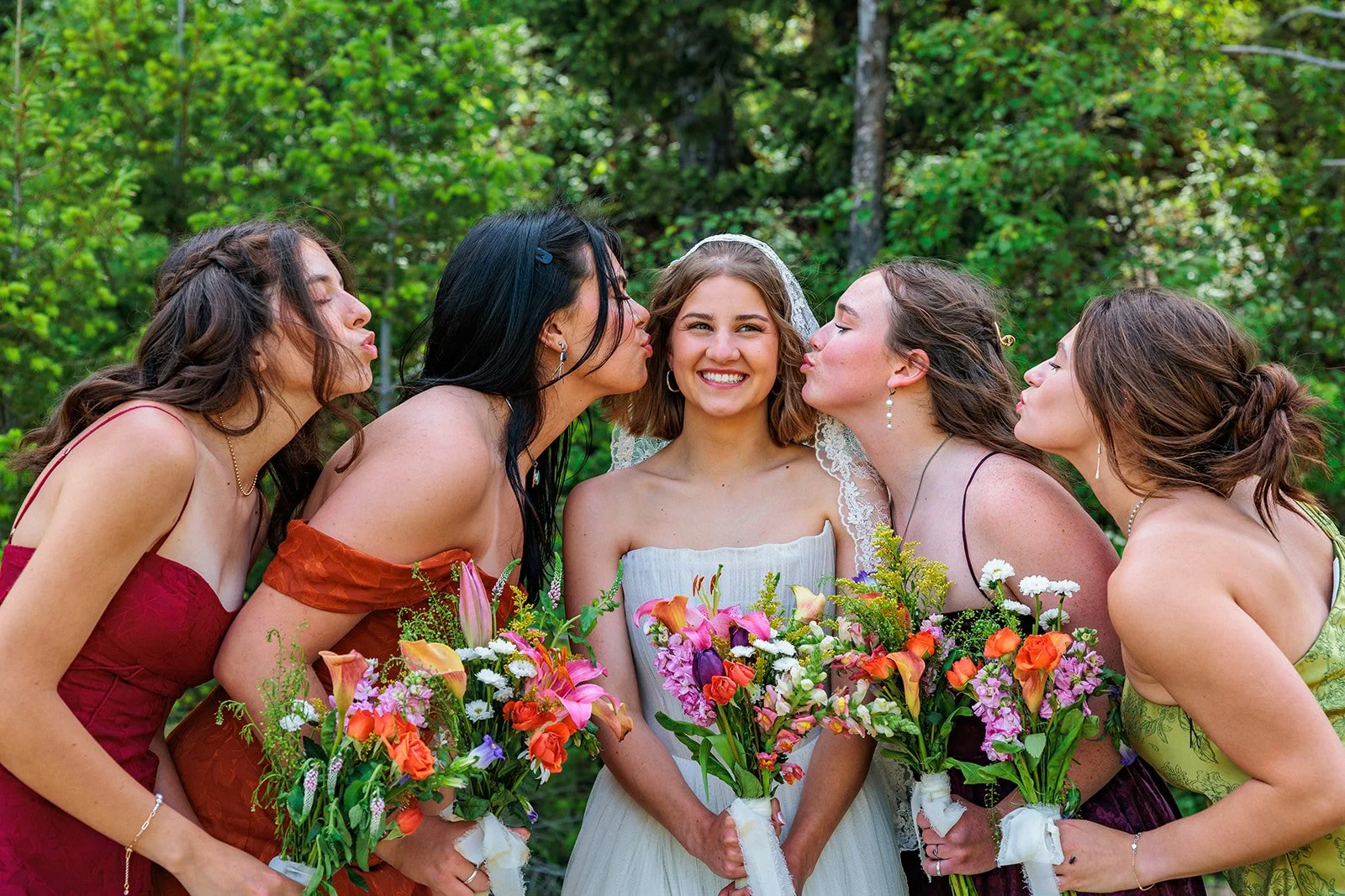 A bride with four bridesmaids, all holding colorful bouquets, outdoors in a green wooded area, with the bridesmaids kissing the bride on her cheeks.