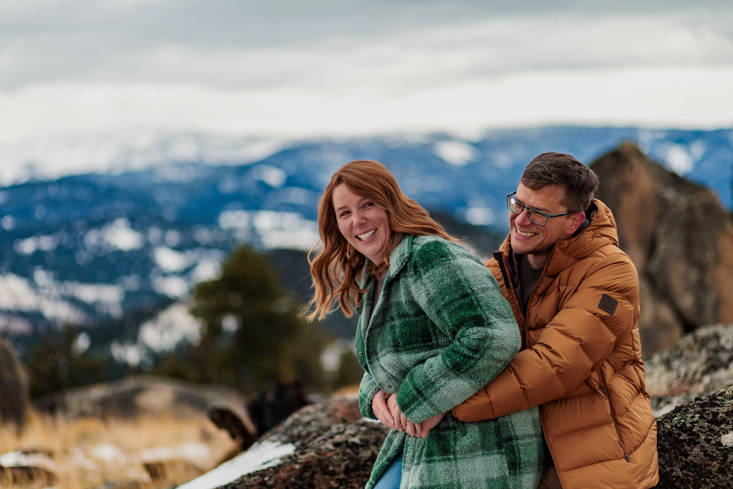 A smiling couple, a woman with red hair wearing a green plaid jacket and a man with glasses wearing an orange puffer jacket, enjoying a moment outdoors in a mountainous area with snow-capped peaks and overcast sky in the background.