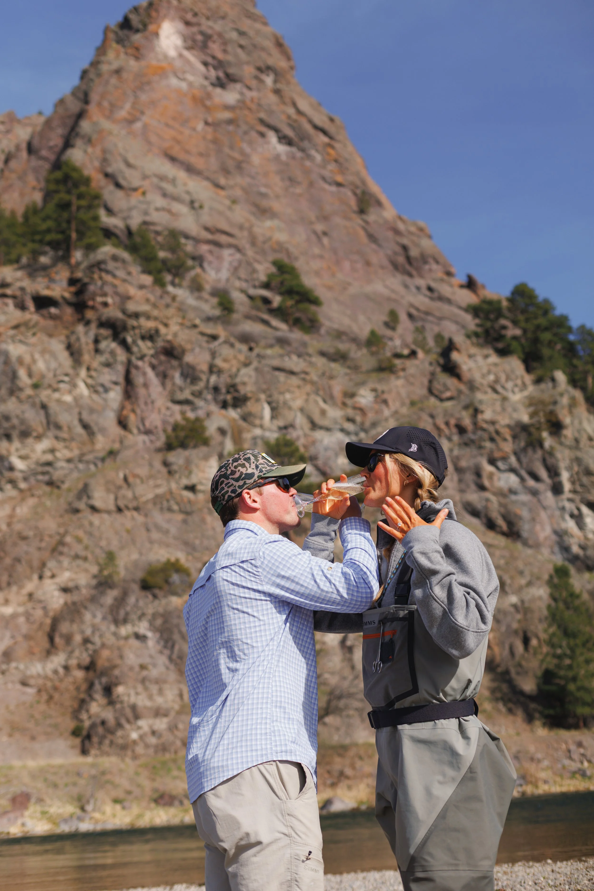 A man and a woman, both wearing outdoor clothing and hats, are drinking wine together outdoors near a rocky hillside.