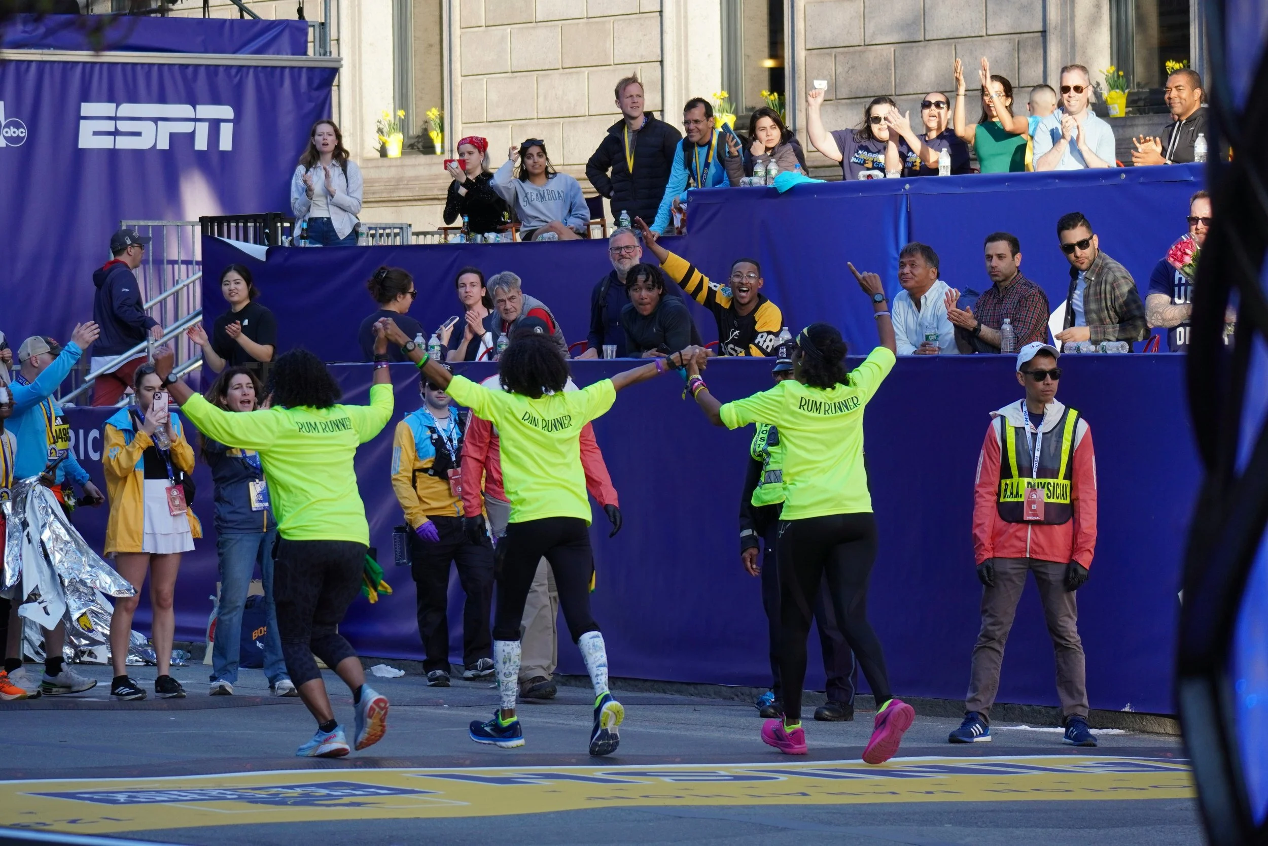 A group of marathon runners wearing neon green shirts crossing the finish line, celebrated by cheering spectators and race staff.