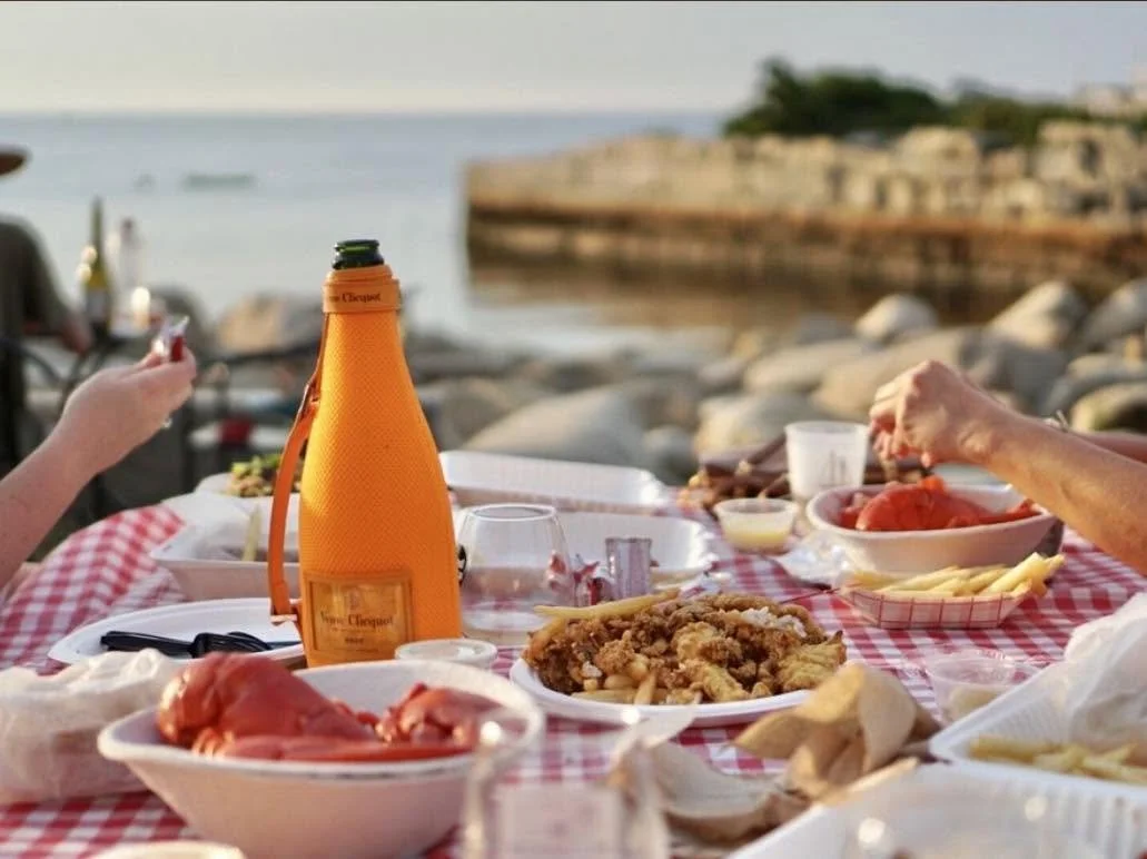 An outdoor seafood dinner by the water at The Lobster Pool in Rockport, Massachusetts, featuring fresh lobster and a bottle of champagne.