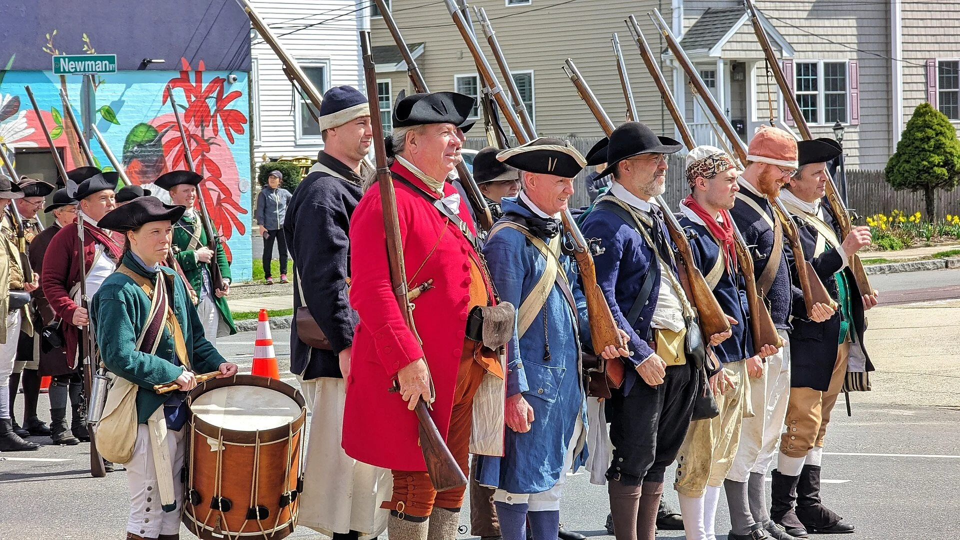 A group of American Revolutionary War re-enactors in period military uniforms and civilian militia attire, carrying muskets and a drum during a public procession or living history event.
