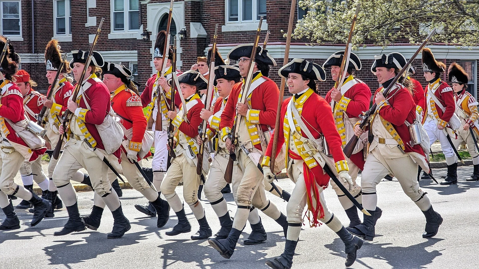 Historical reenactors dressed as British Redcoats marching in a parade, likely during a Patriot's Day