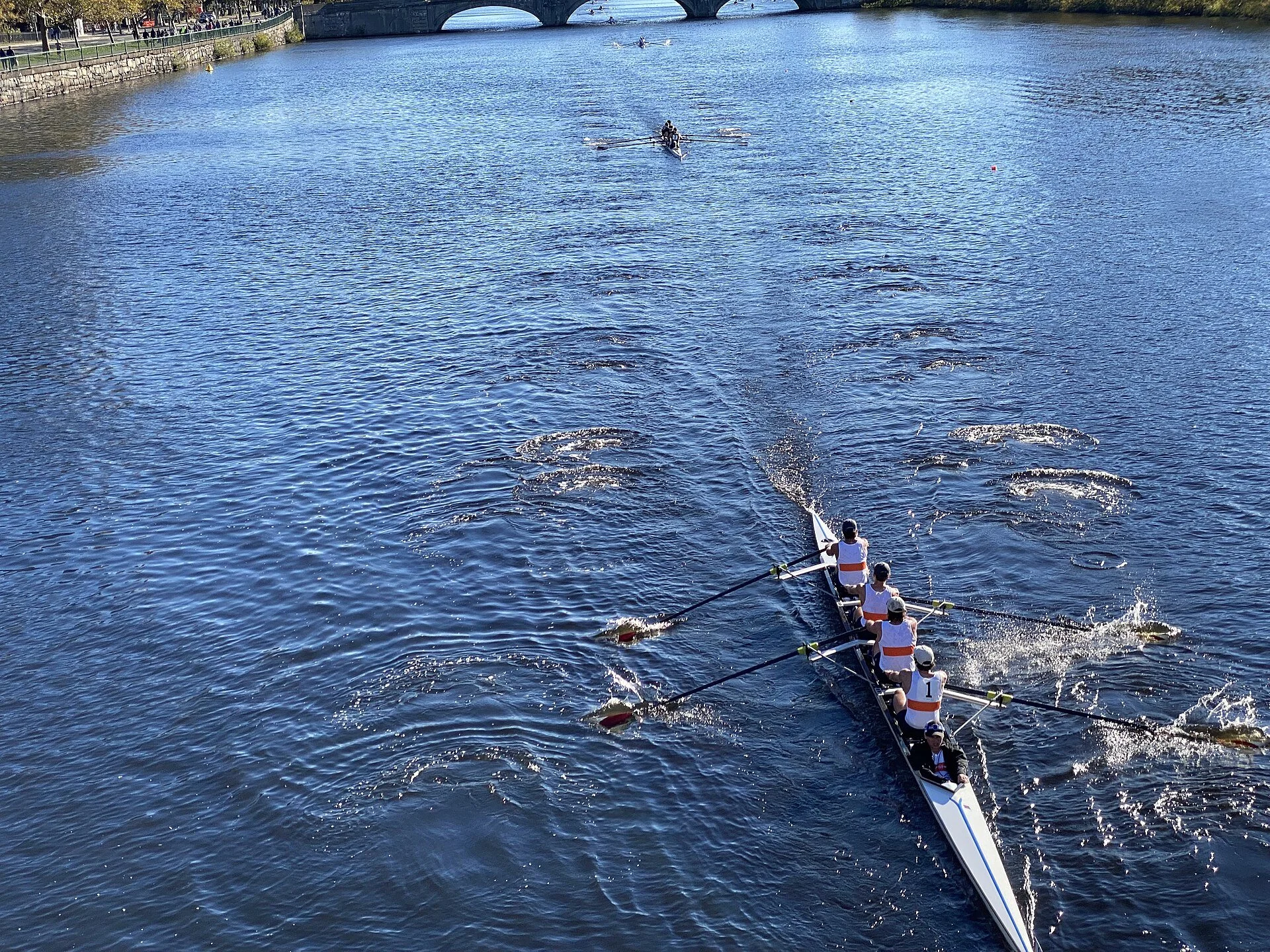 Crew navigating a narrow river course, viewed from an overhead bridge with concentric ripple patterns trailing behind their oars.