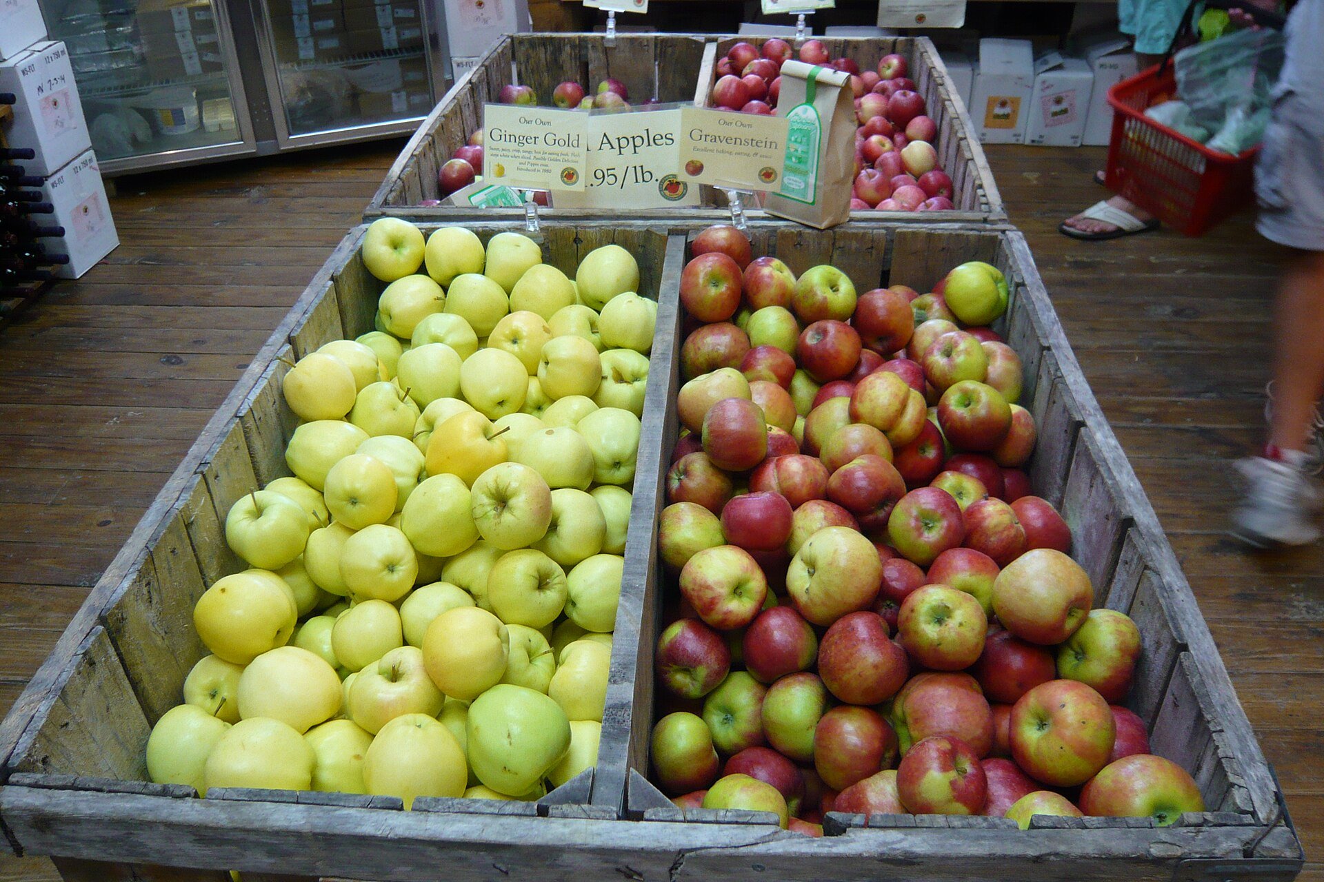 Two wooden crates of fresh apples in a market, with the left crate containing pale greenish-yellow Ginger Gold apples and the right crate featuring red-blushed Gravenstein apples