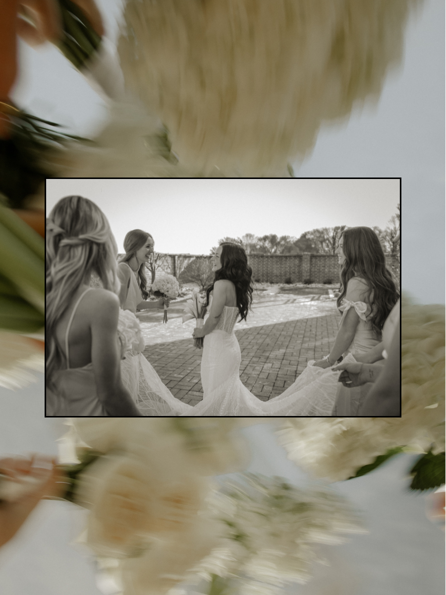 A black and white photo of a bride in a wedding dress holding a bouquet, surrounded by her bridesmaids, outside on a brick patio with a brick wall and trees in the background.