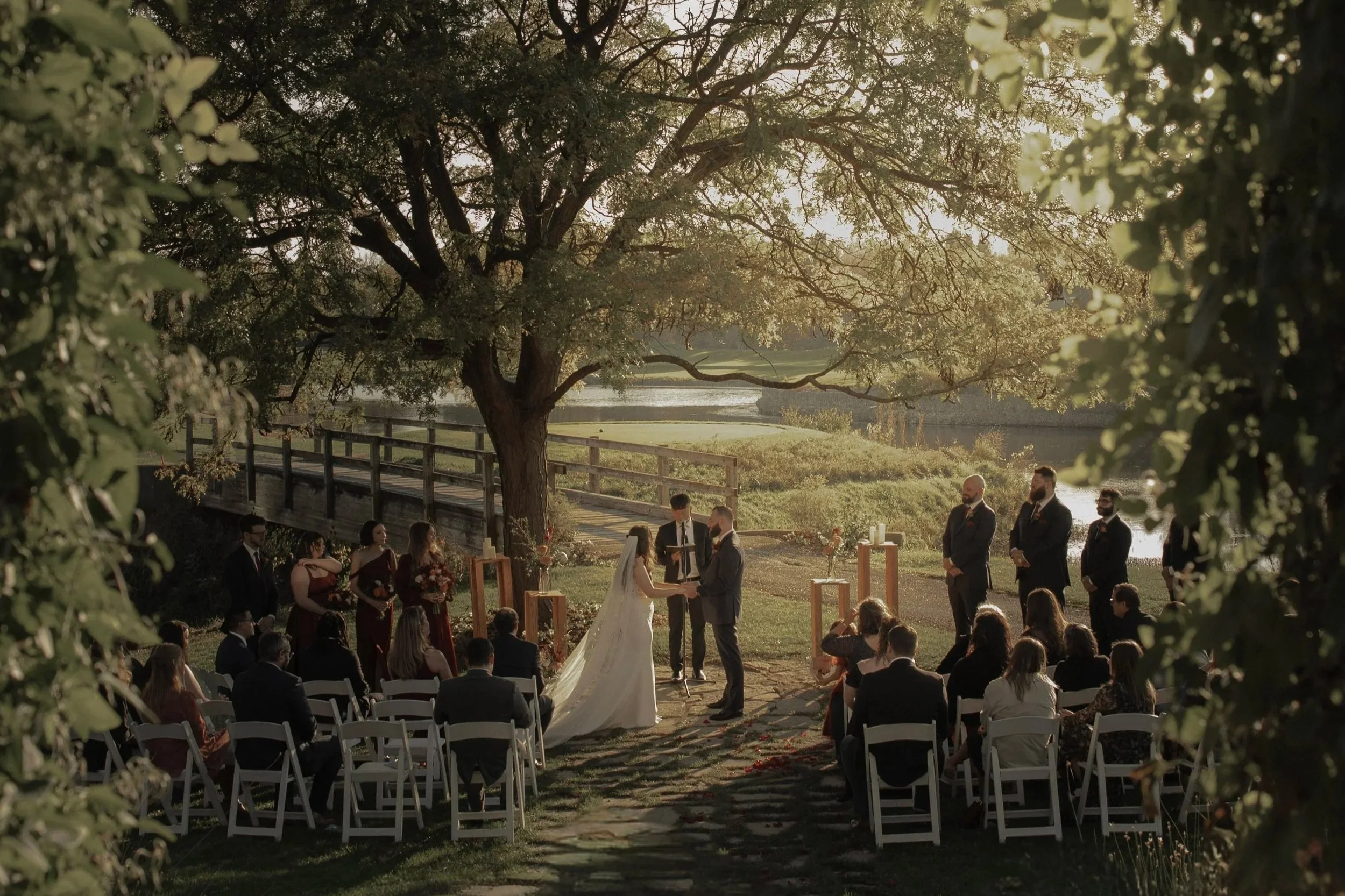 Outdoor wedding ceremony under a large tree with guests seated in white chairs, the bride and groom holding hands, in a scenic natural setting during sunset.