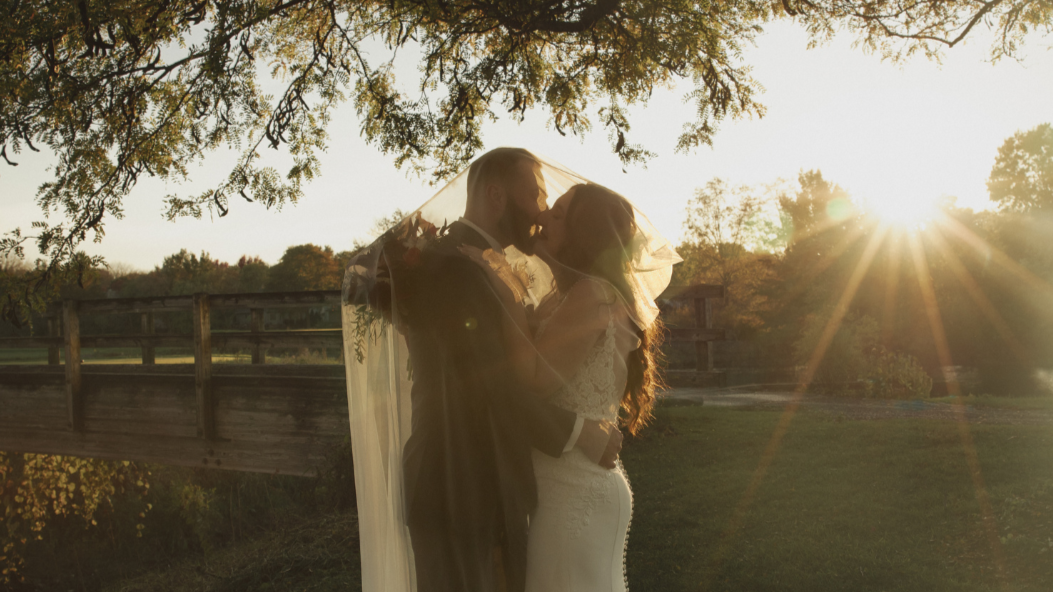A bride and groom share a kiss outdoors during sunset, with a wooden bridge and trees in the background.
