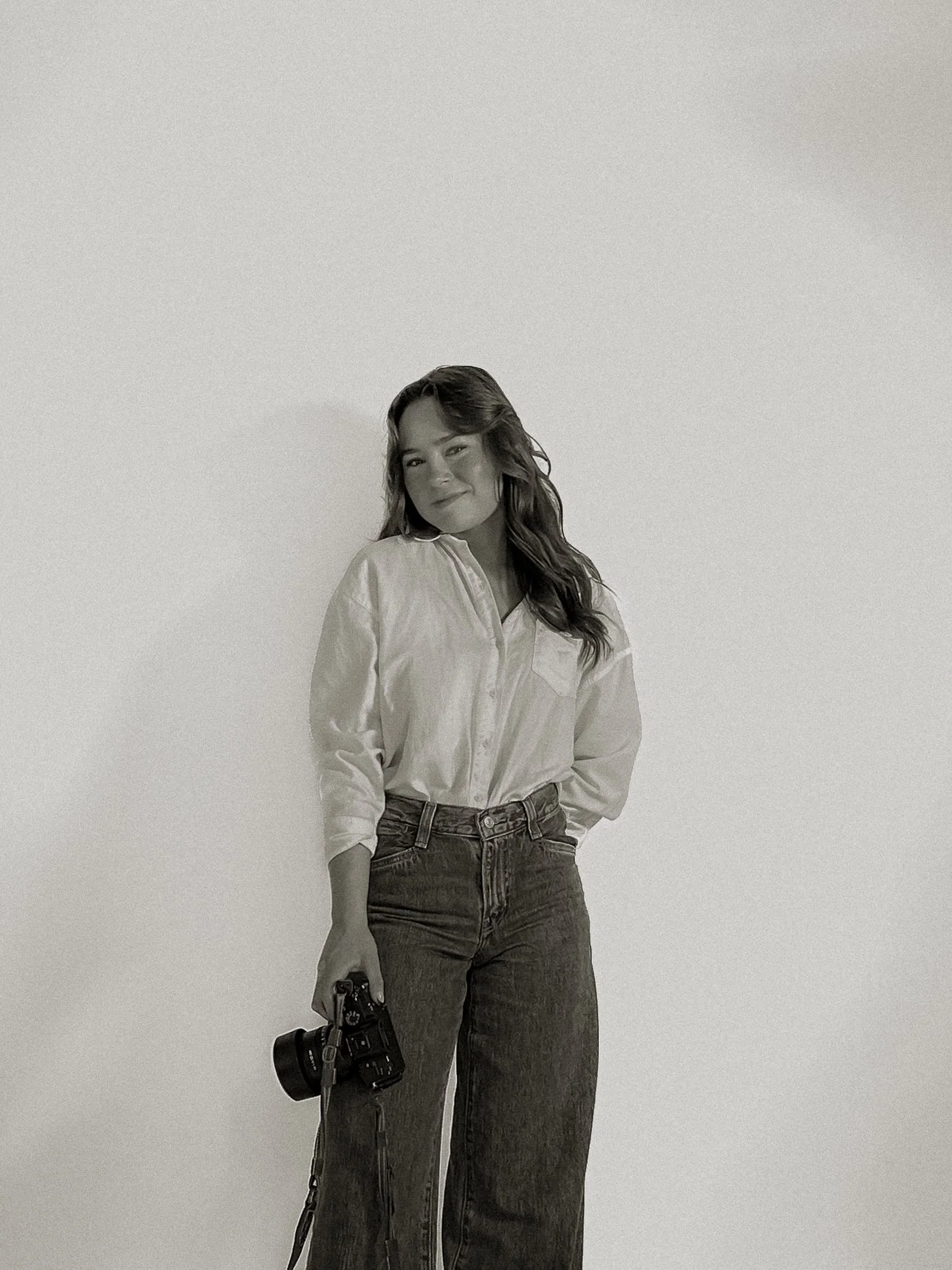 A woman with wavy hair, wearing a white button-up shirt and high-waisted jeans, holding a camera in her right hand, standing against a plain wall.