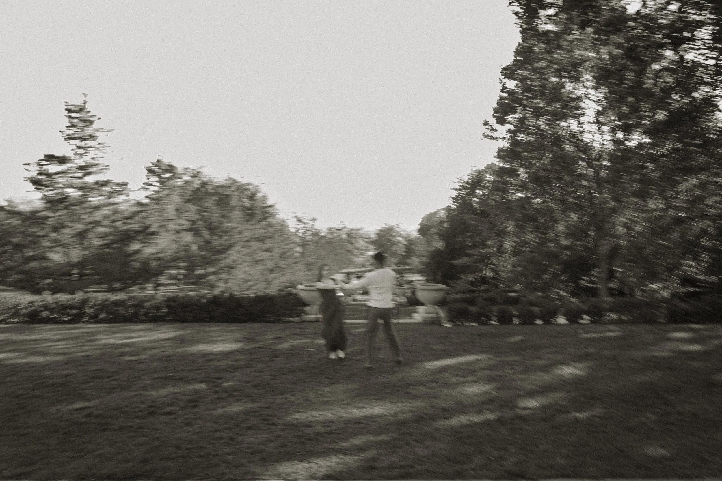 A blurred black-and-white photo of a man and woman dancing outdoors in a park-like setting with trees and a row of boats in the background.