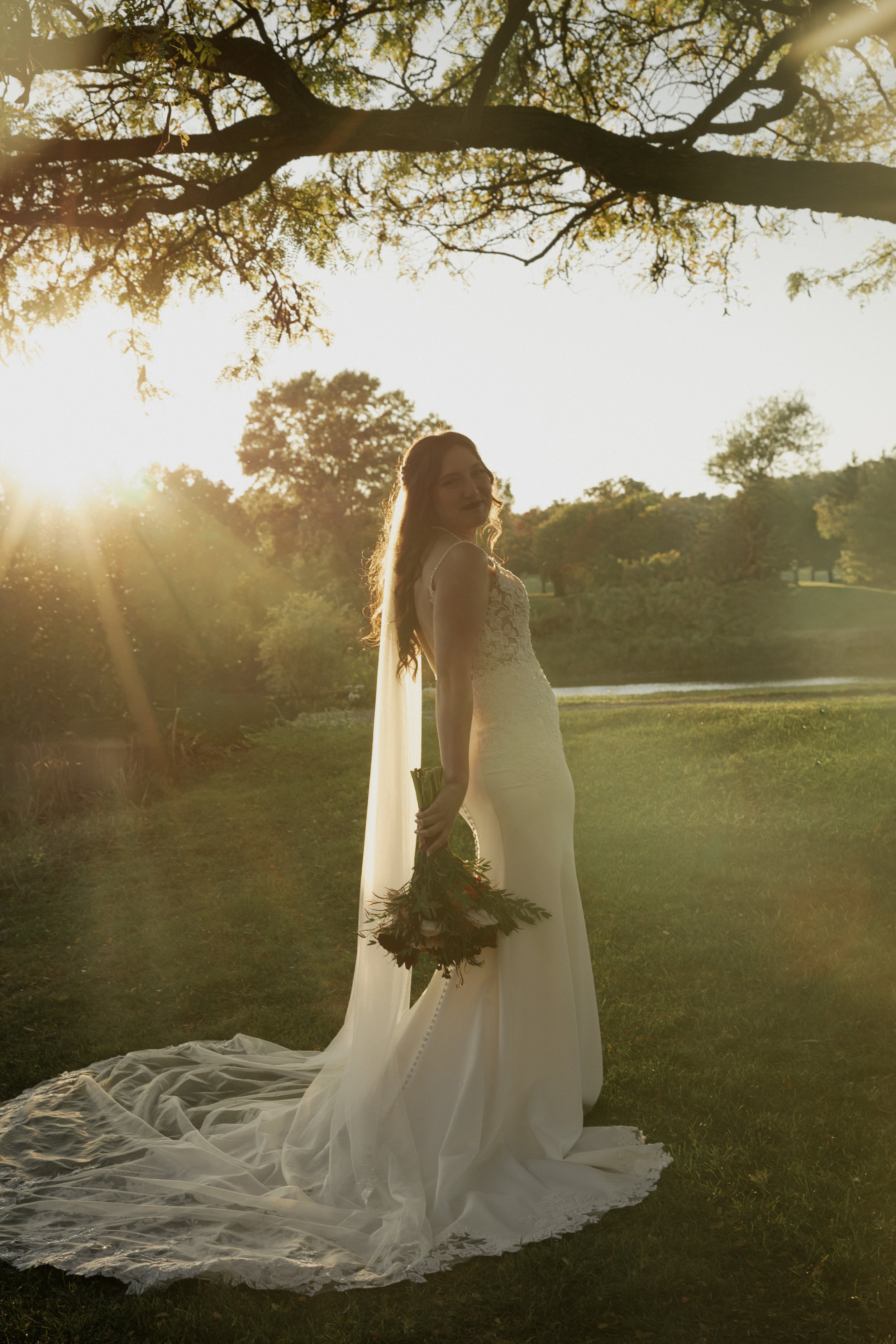 A woman in a wedding dress standing outdoors at sunset, holding a bouquet of flowers, with trees and a lake in the background.