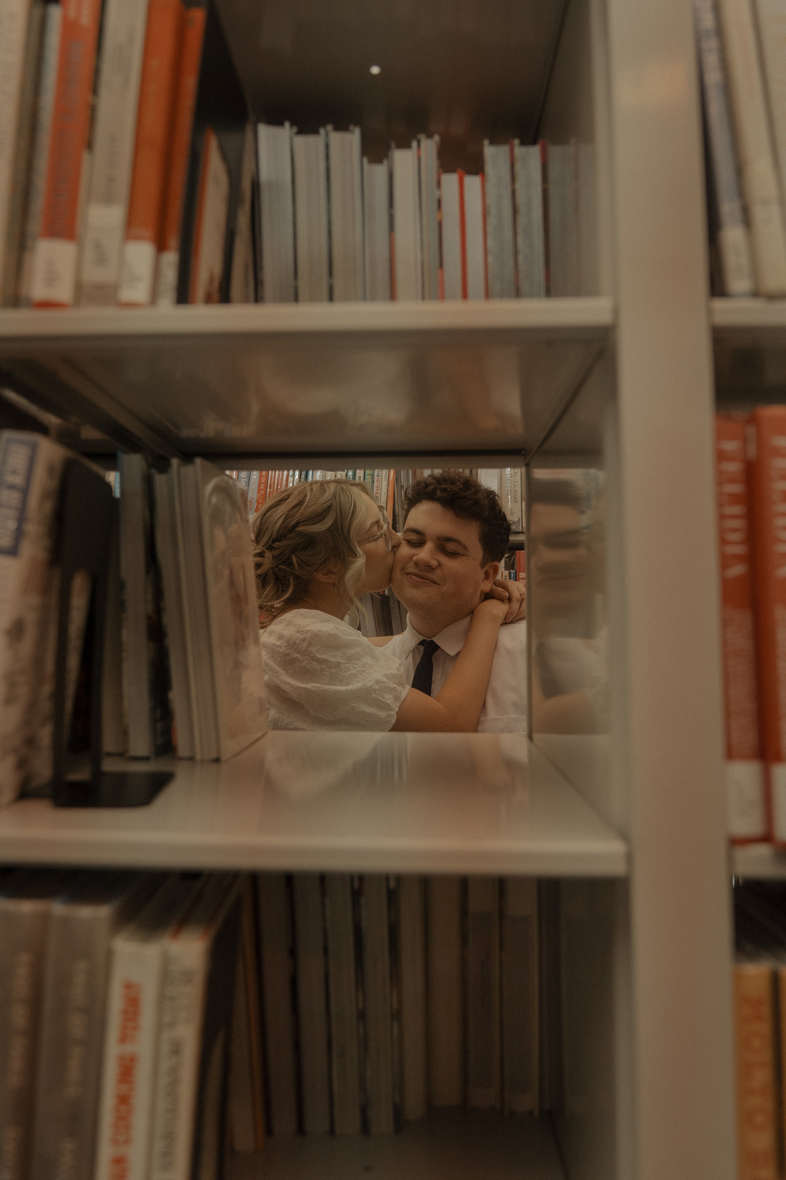 A woman giving a kiss on the cheek to a young man inside a library, seen through a gap between bookshelves.