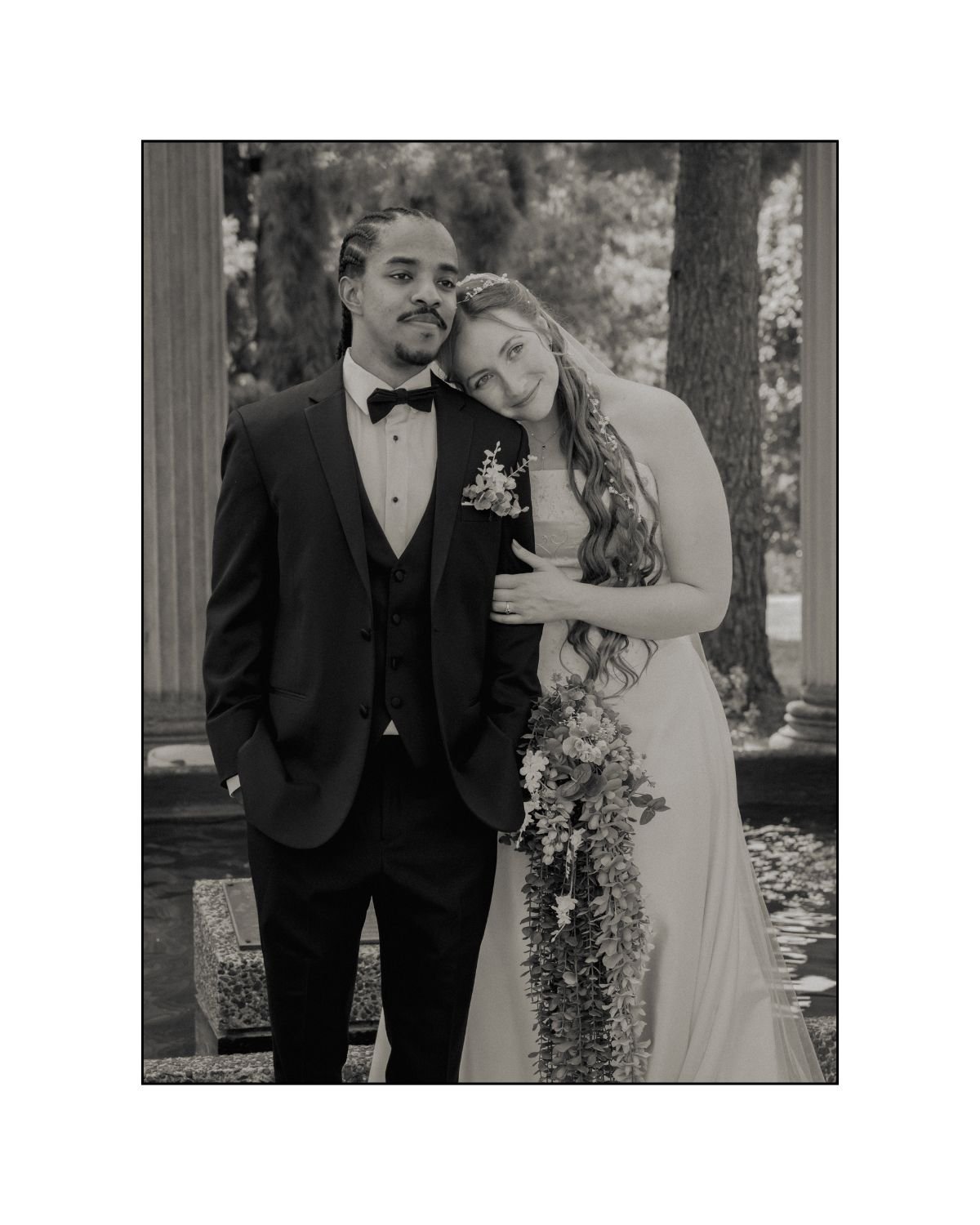 Black and white photo of a newlywed couple standing outdoors. The groom is wearing a tuxedo with a bow tie and has his hands in his pockets. The bride is leaning her head on his shoulder, holding a long bouquet of flowers, and wearing a wedding dress