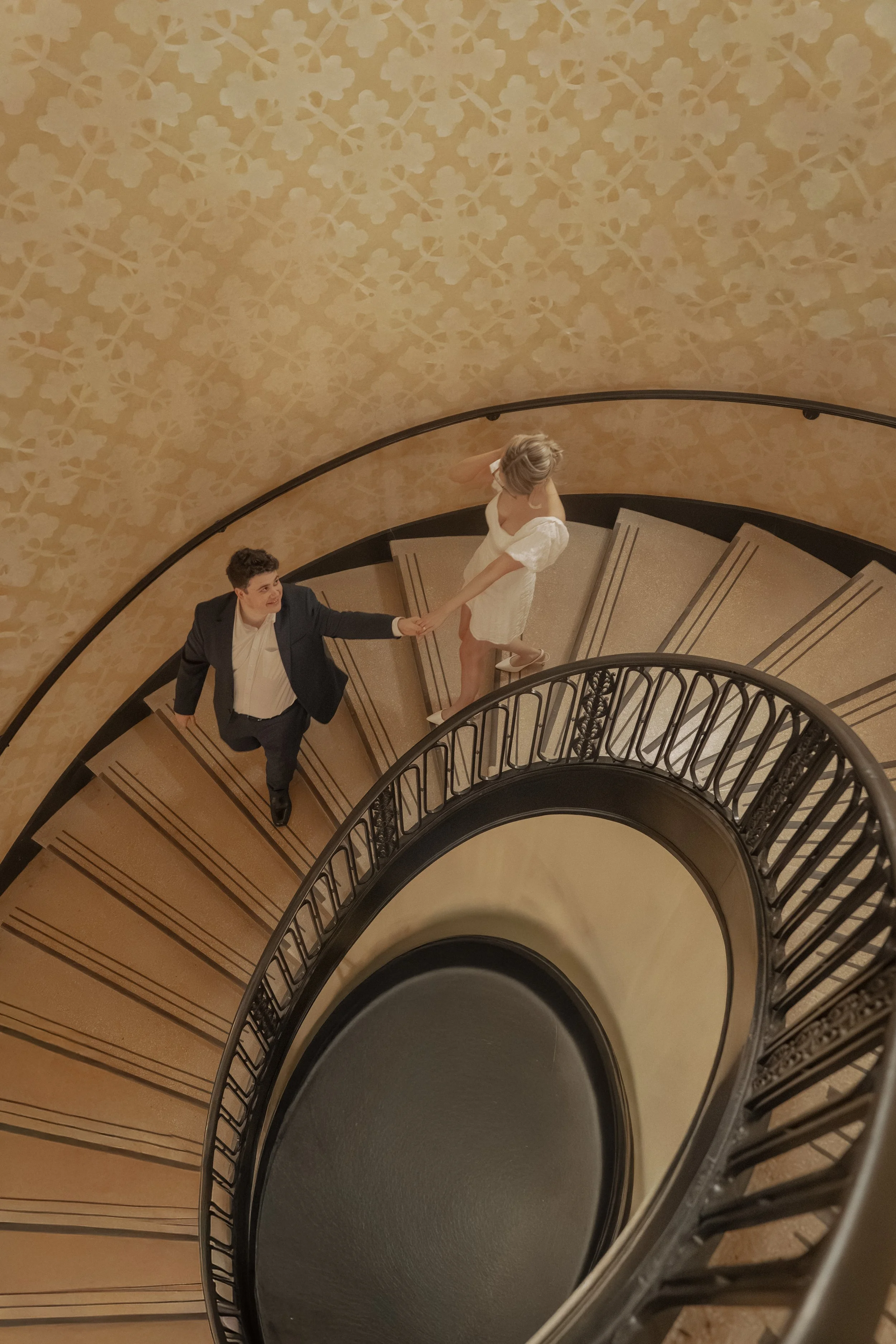 A man dressed in a suit is holding hands with a woman in a white dress on a spiral staircase, viewed from above.