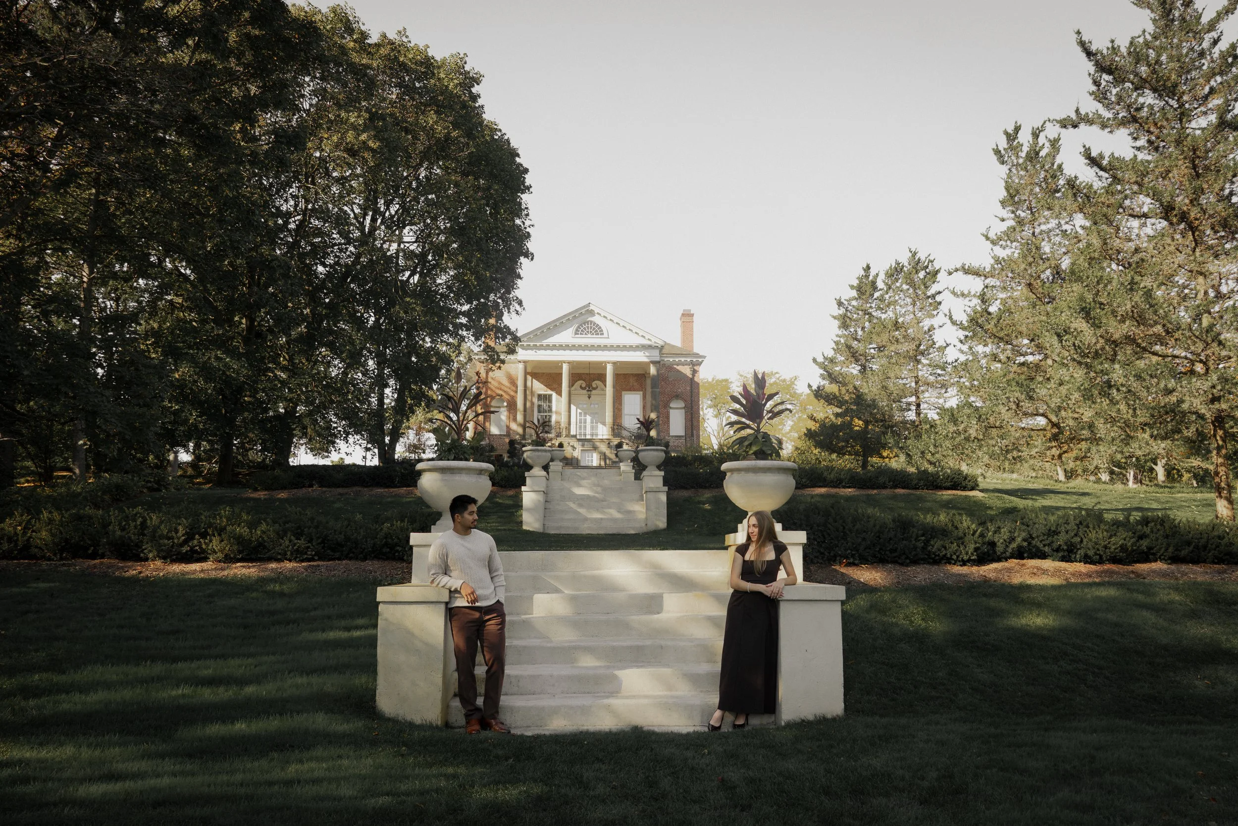 A man and woman standing on the steps of a large mansion in a garden with lush trees and potted plants.