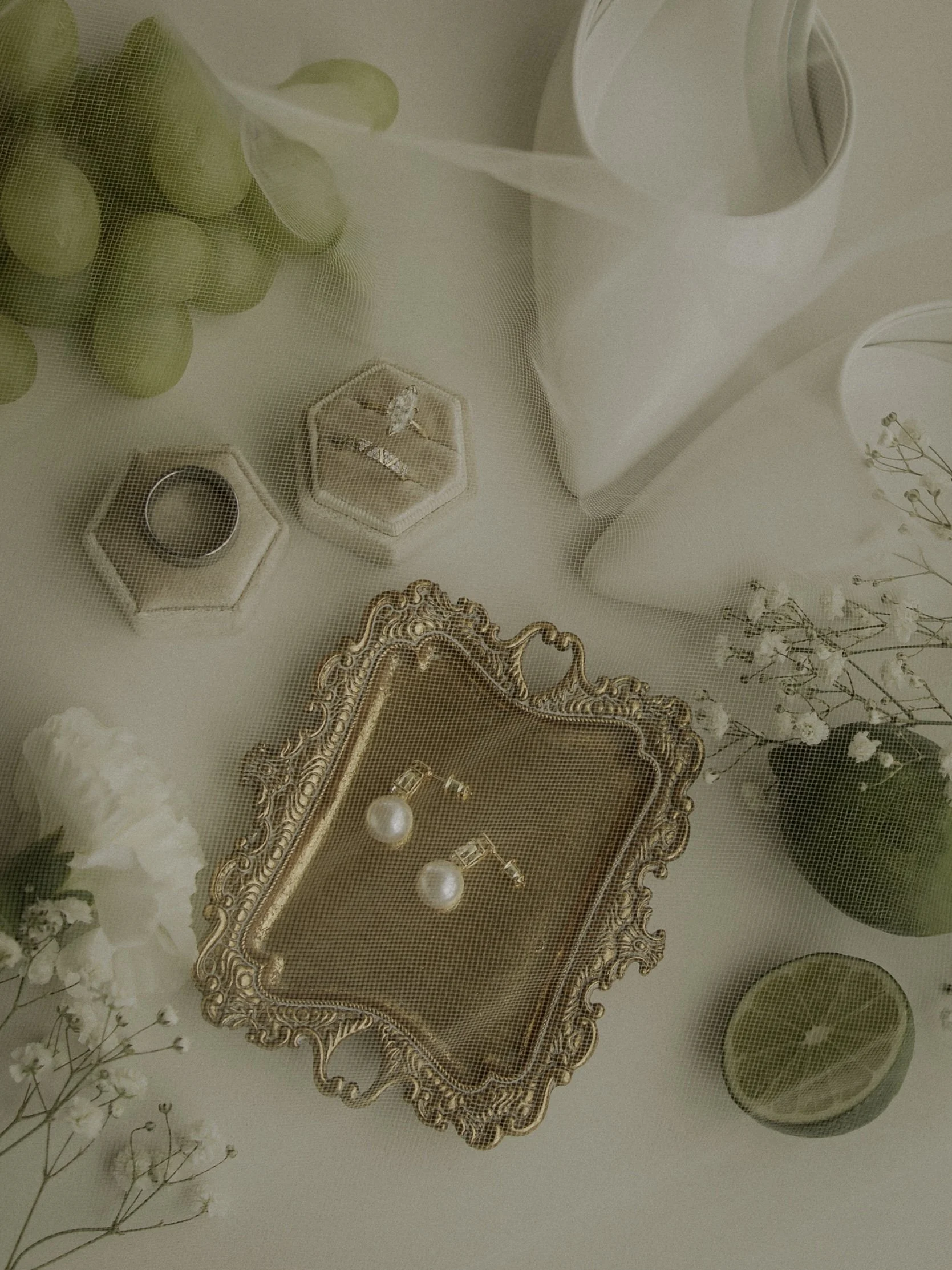 Jewelry display with pearl earrings, rings, and decorative elements on a vintage gold tray, surrounded by green grapes, a white pitcher, lime halves, and white flowers.