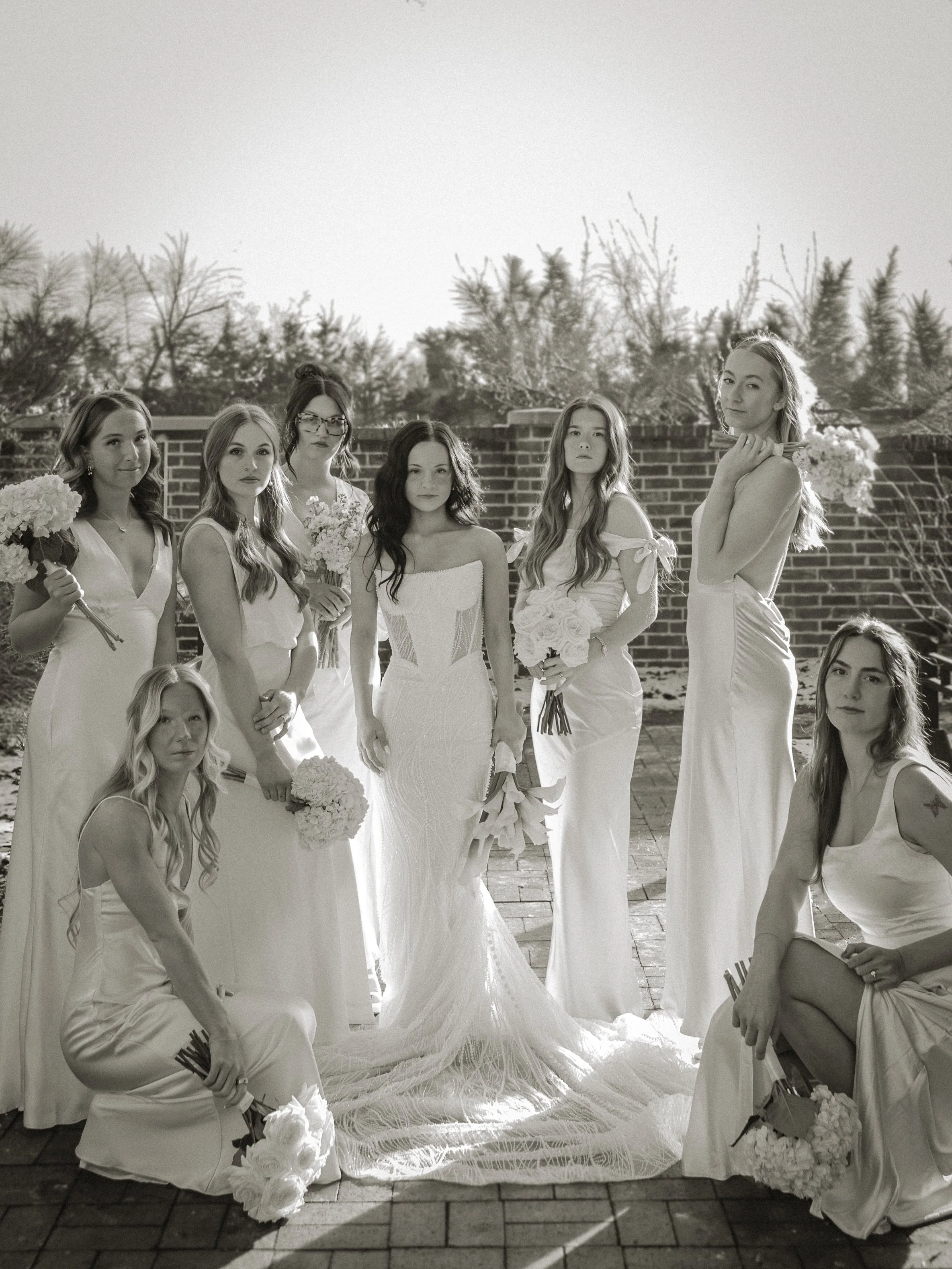 Black and white photo of a group of women, likely bridesmaids, standing outdoors on a patio with a brick wall and trees in the background. They are wearing elegant dresses and holding bouquets of flowers.